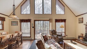 A spacious living room with a high ceiling, wooden furniture, and large windows. - Hot Tub + Sauna + Deck + Firepit + Beach Access + Near Dining Grateful Escape by Avantstay (Lake Harmony)