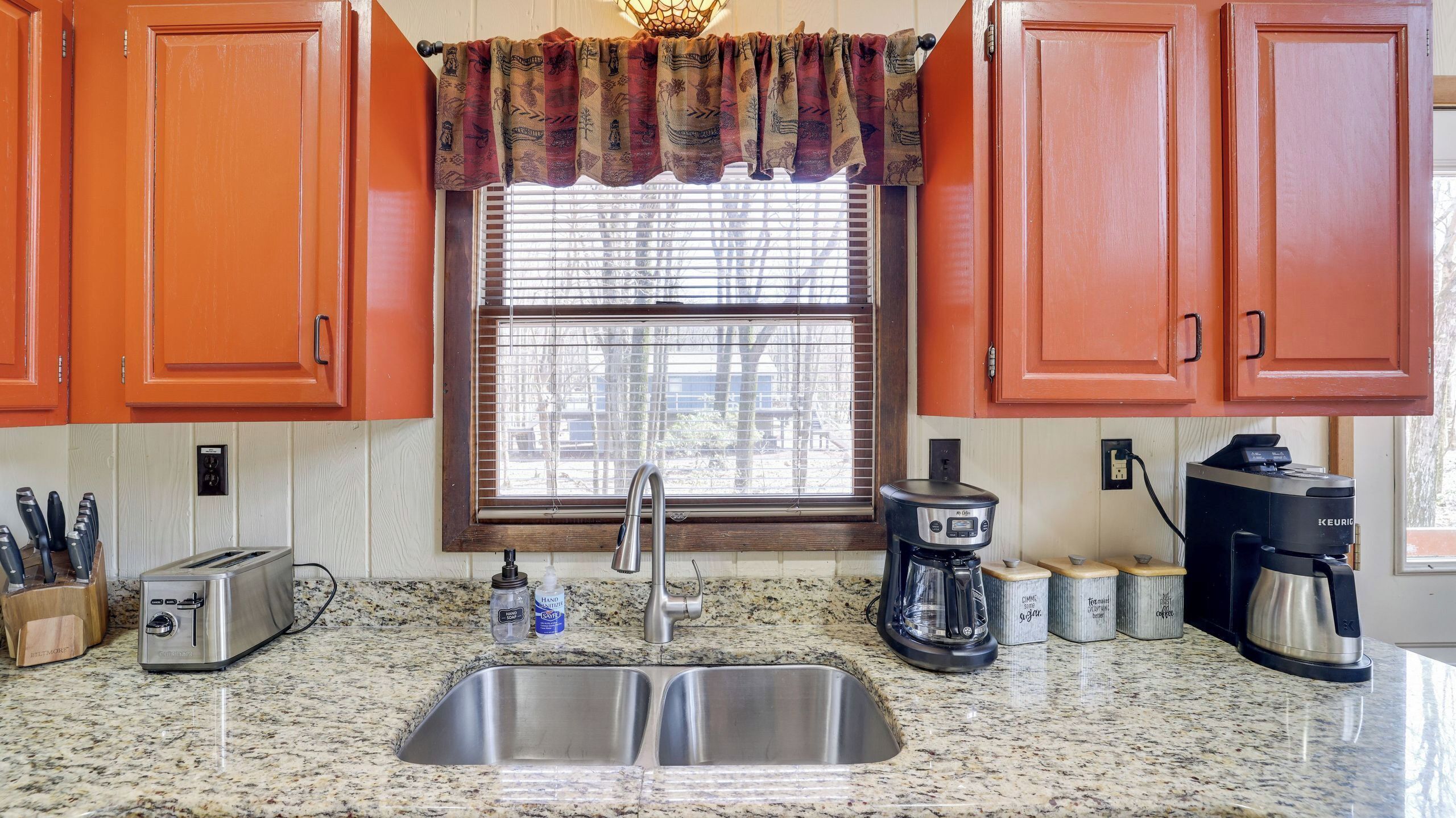 A kitchen with a stainless steel sink, a toaster, a coffee maker, and a window with blinds.