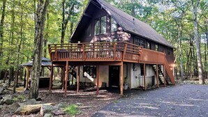 A cabin with a wooden deck and a large window, surrounded by trees. - Hot Tub + Sauna + Deck + Firepit + Beach Access + Near Dining Grateful Escape by Avantstay (Lake Harmony)
