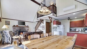 A spacious living room with a large wooden dining table, comfortable seating, a fireplace, and a staircase leading to an upper level. - Hot Tub + Sauna + Deck + Firepit + Beach Access + Near Dining Grateful Escape by Avantstay (Lake Harmony)