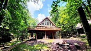 A cabin with a wooden deck, surrounded by trees. - Woodland Oasis by Avantstay Hot Tub, Firepit, Walk to Lake (Lake Harmony)