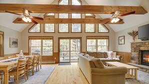 A spacious living room with wooden floors, a large sofa, a dining table with chairs, and a fireplace. - Woodland Oasis by Avantstay Hot Tub, Firepit, Walk to Lake (Lake Harmony)