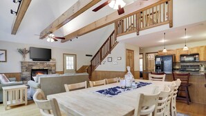 A spacious living area with a fireplace, wooden dining table, and a kitchen with modern appliances. - Woodland Oasis by Avantstay Hot Tub, Firepit, Walk to Lake (Lake Harmony)