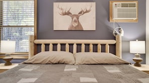 A bedroom with a wooden headboard, two pillows, a patterned bedspread, a wall-mounted air conditioner, and a window with blinds.
