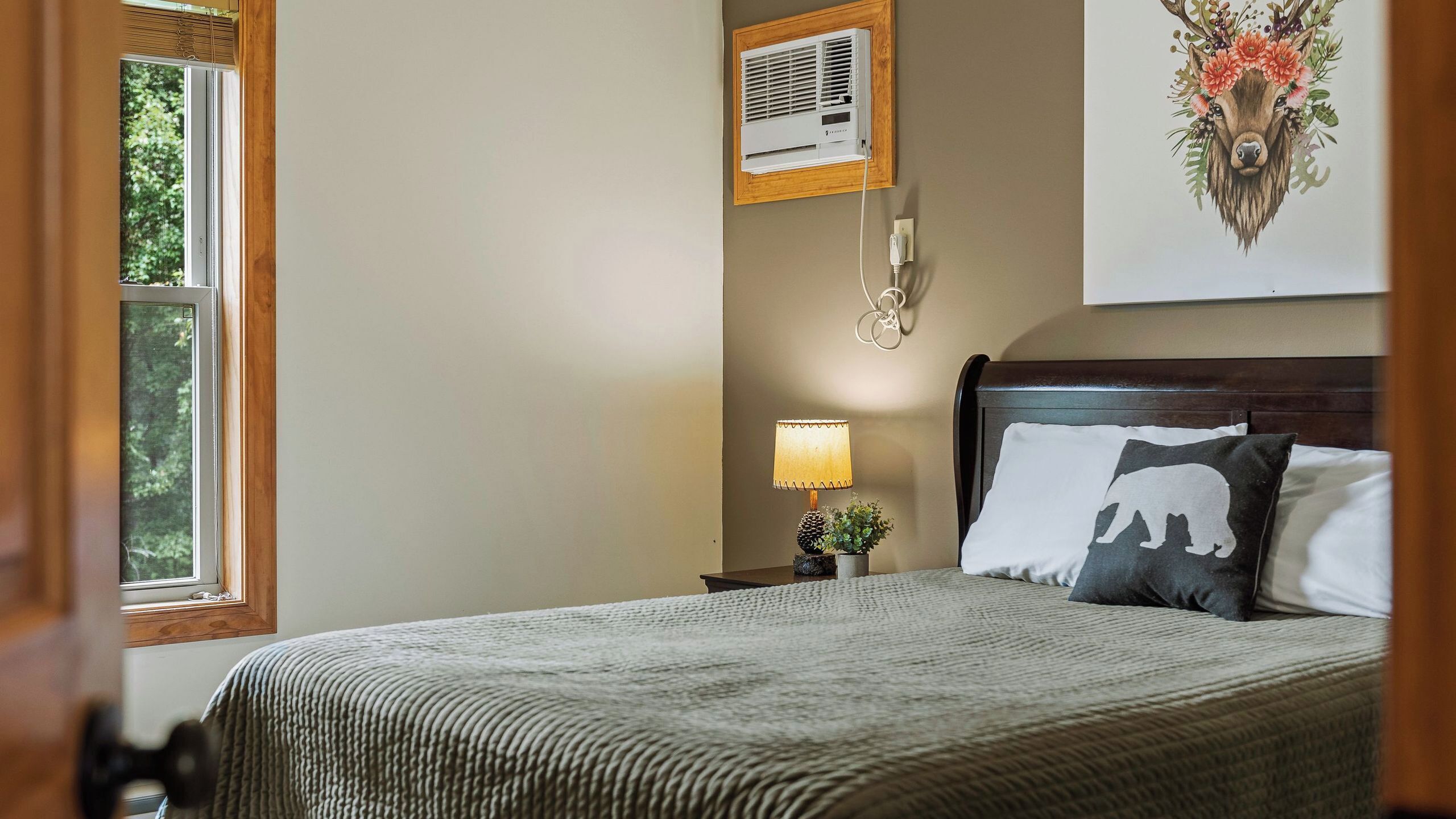 A bedroom with a bed, a nightstand, a lamp, a wall-mounted air conditioner, and a window with a view of trees.