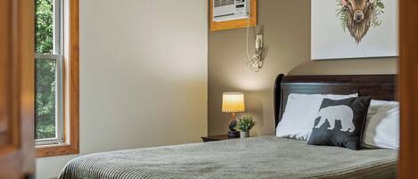 A bedroom with a bed, a nightstand, a lamp, a wall-mounted air conditioner, and a window with a view of trees.
