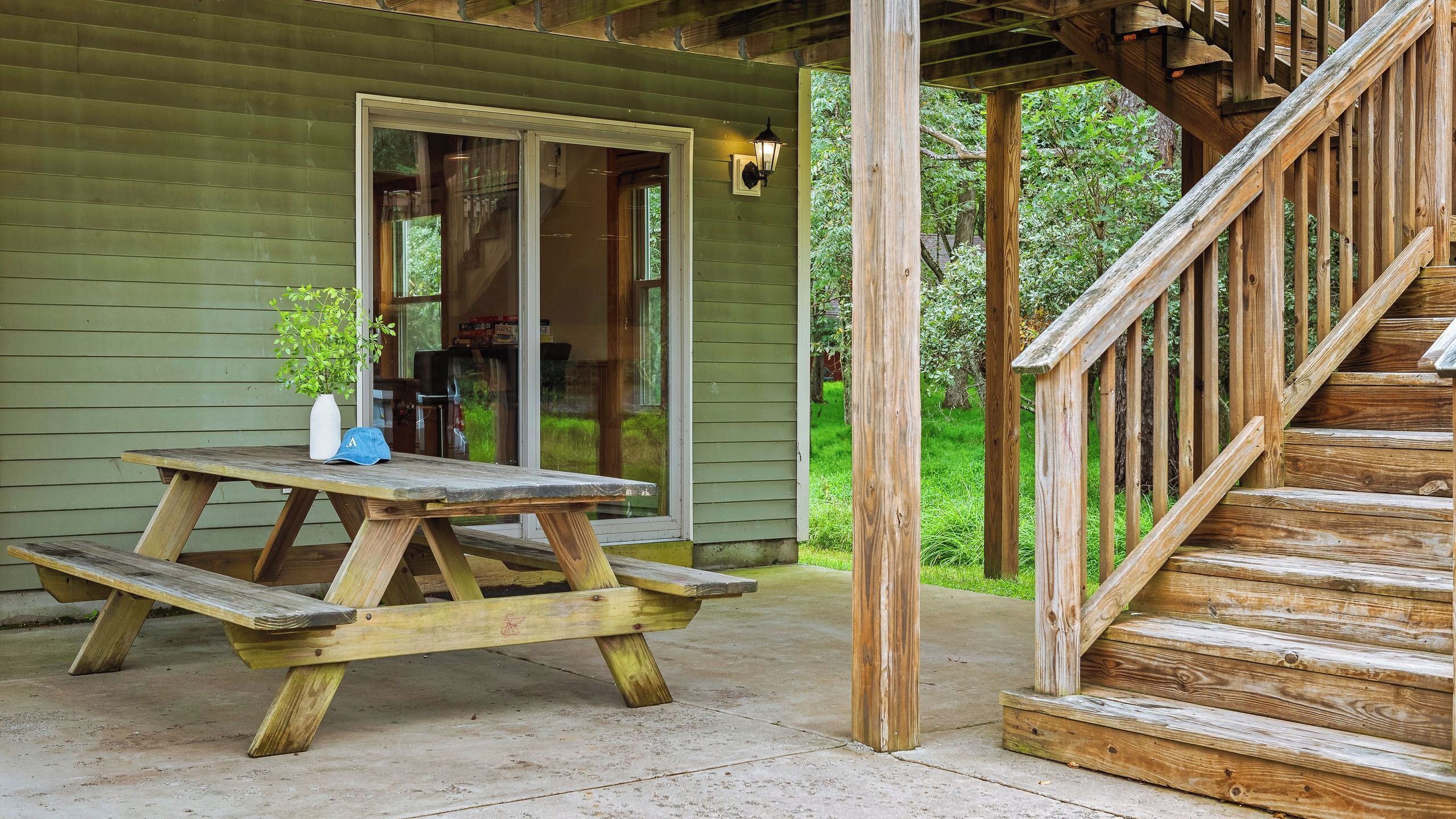 A wooden picnic table with a white vase and a blue cap, a wooden staircase, and a green building with a sliding glass door.