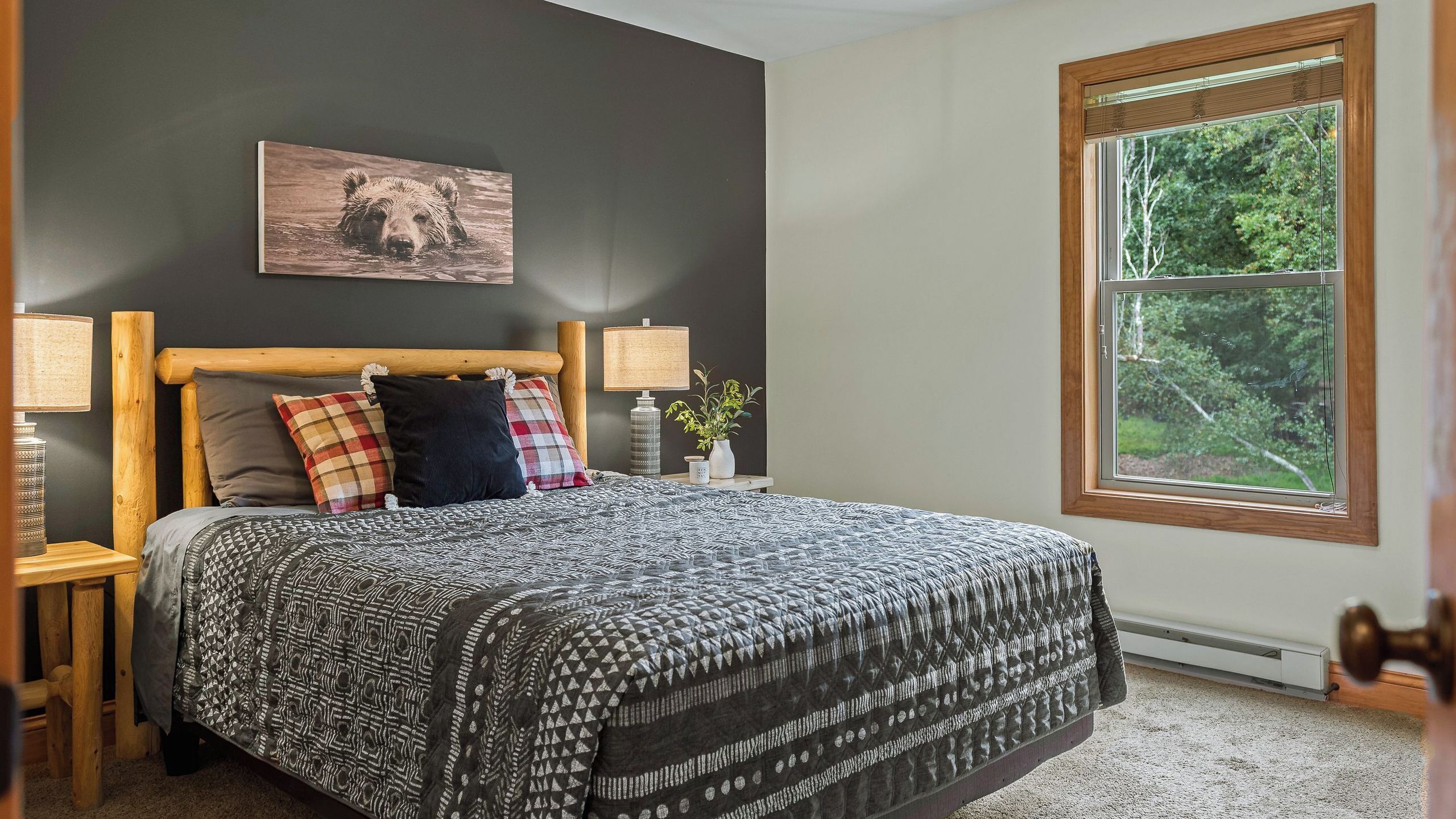 A bedroom with a large bed, a headboard, a bedside table with a lamp, a wall-mounted picture, and a window with a view of trees.