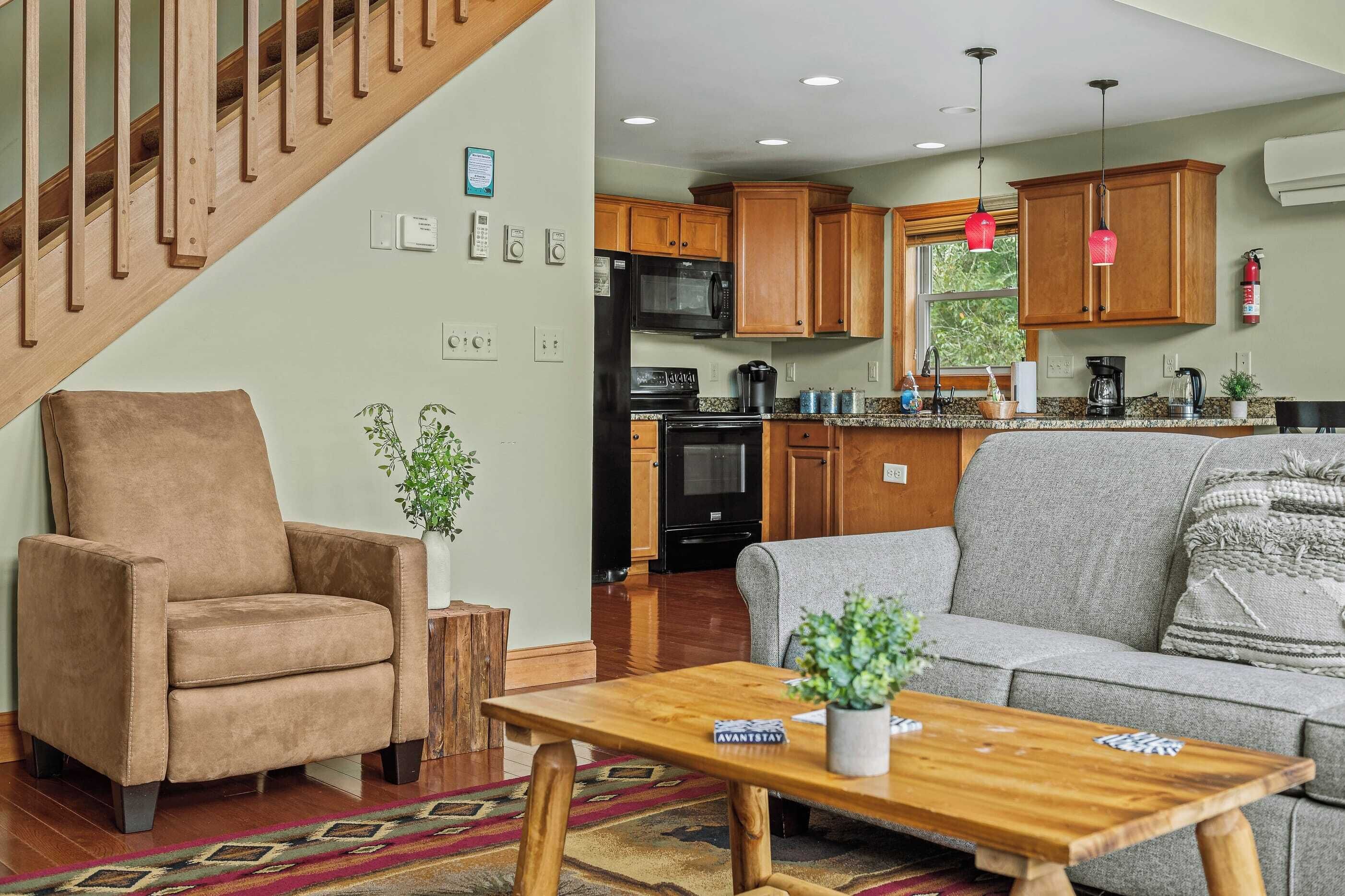A living room with a sofa, armchair, and coffee table, a kitchen with wooden cabinets and appliances, and a staircase.