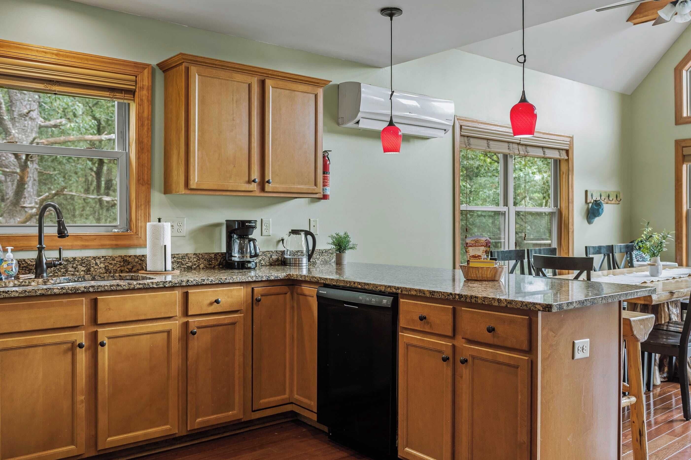 A kitchen with wooden cabinets, a dishwasher, granite countertops, and a window overlooking greenery.
