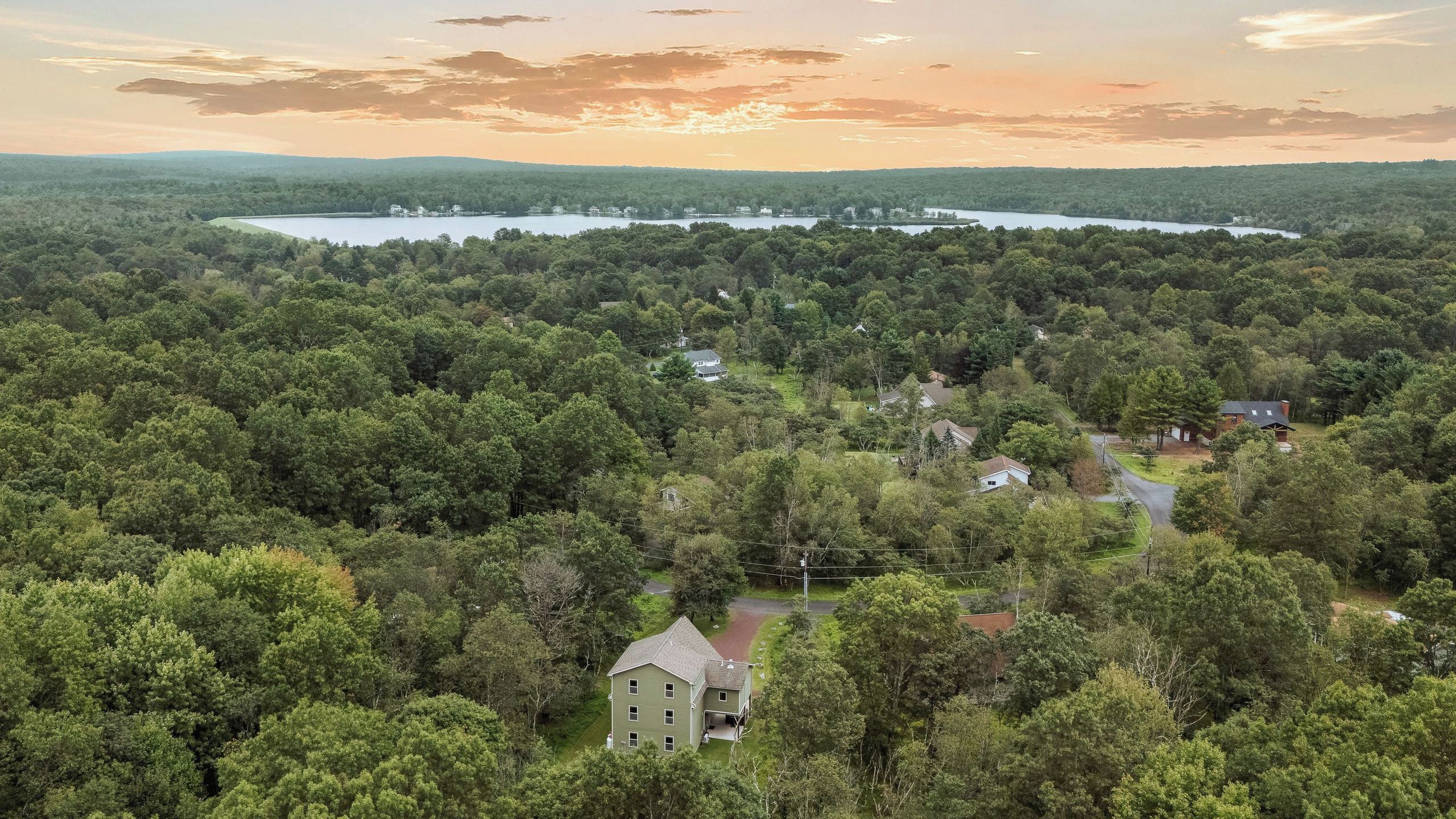 A large house surrounded by dense forest, with a lake and hills in the background.