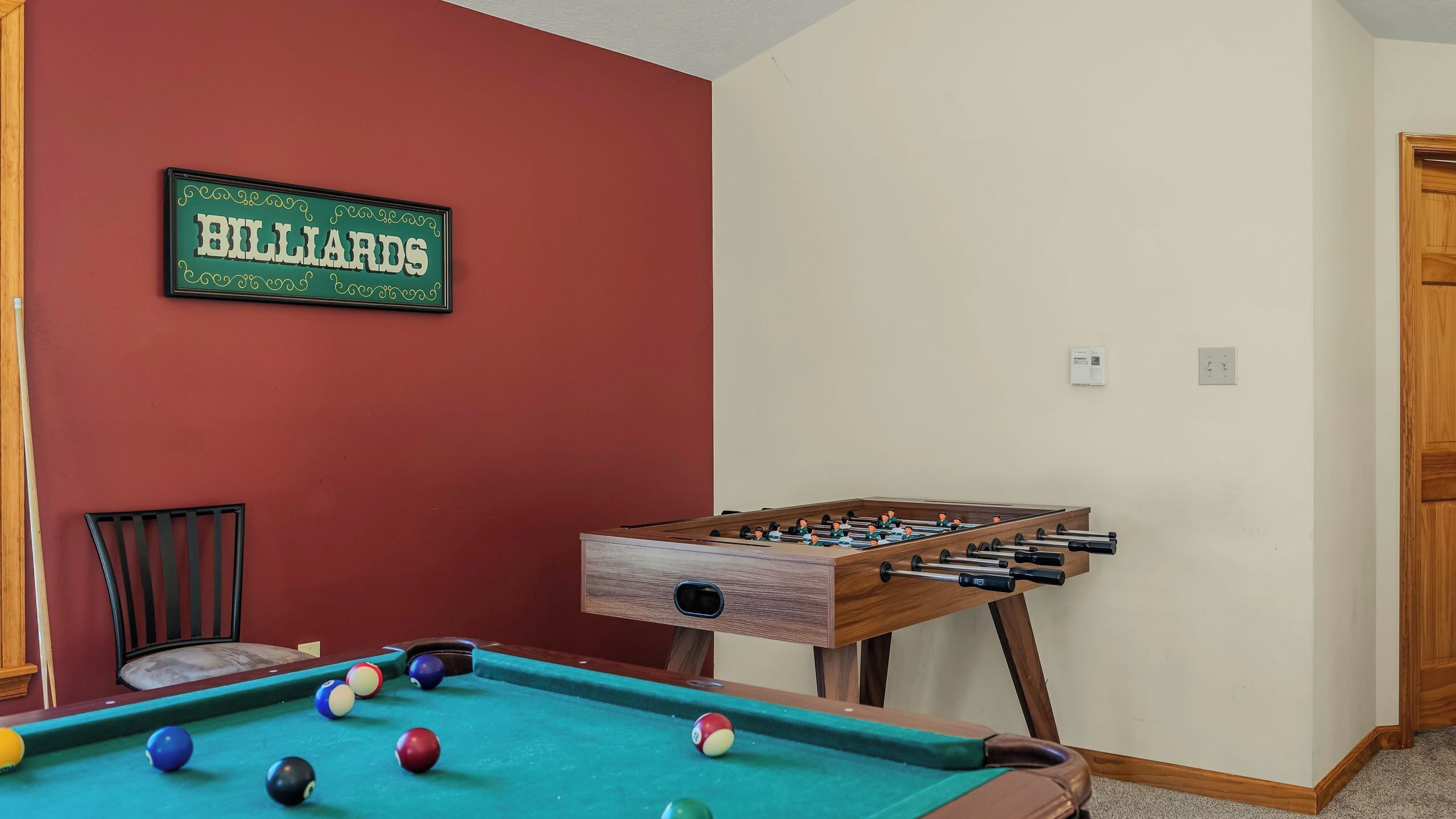 Un tavolo da biliardo con le palle, un tavolo da calcio, una insegna con scritto biliardo, una sedia e una porta di legno.