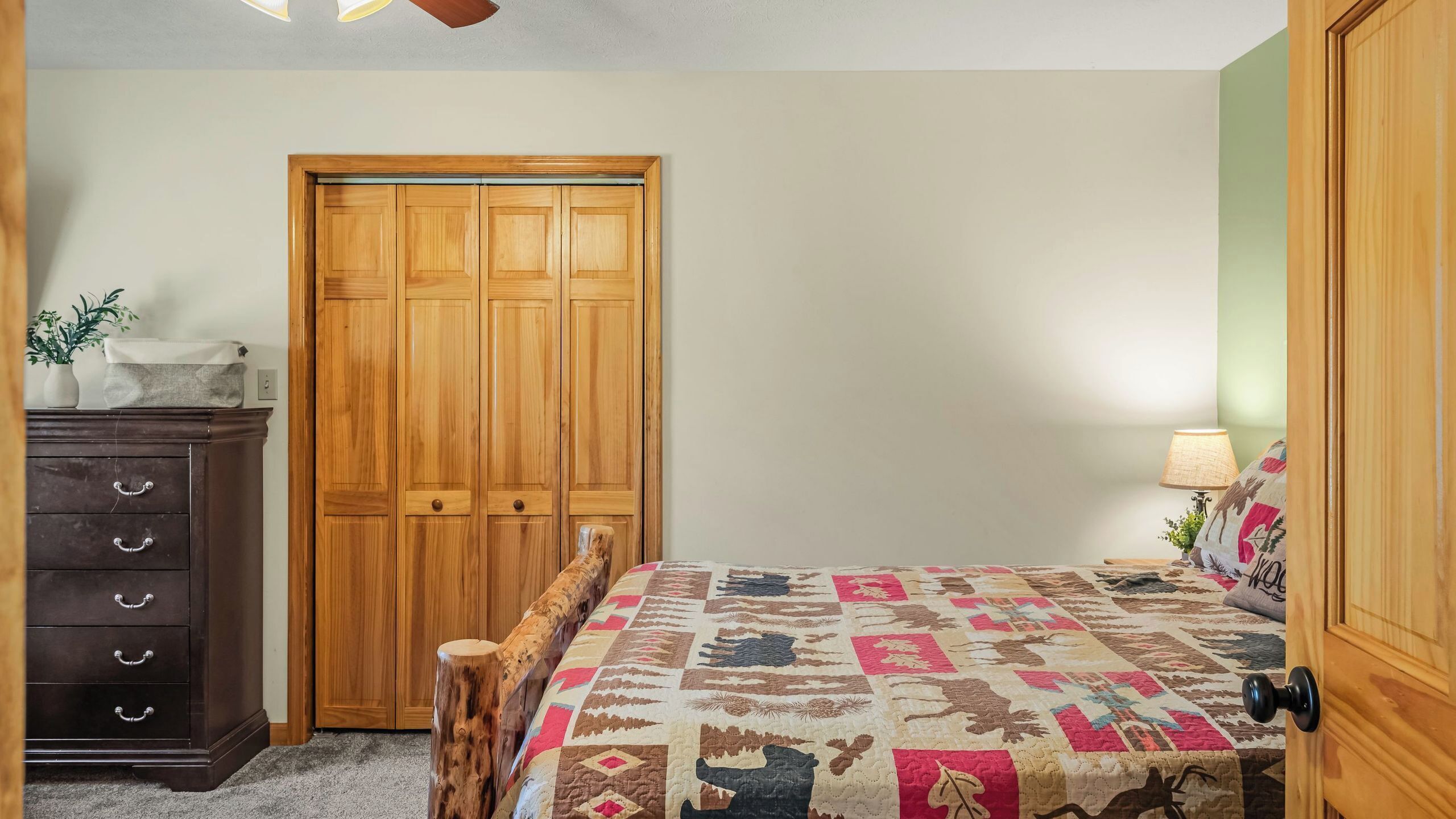 A bedroom with a wooden wardrobe, a dark dresser, a bed with a patterned quilt, and a nightstand with a lamp.