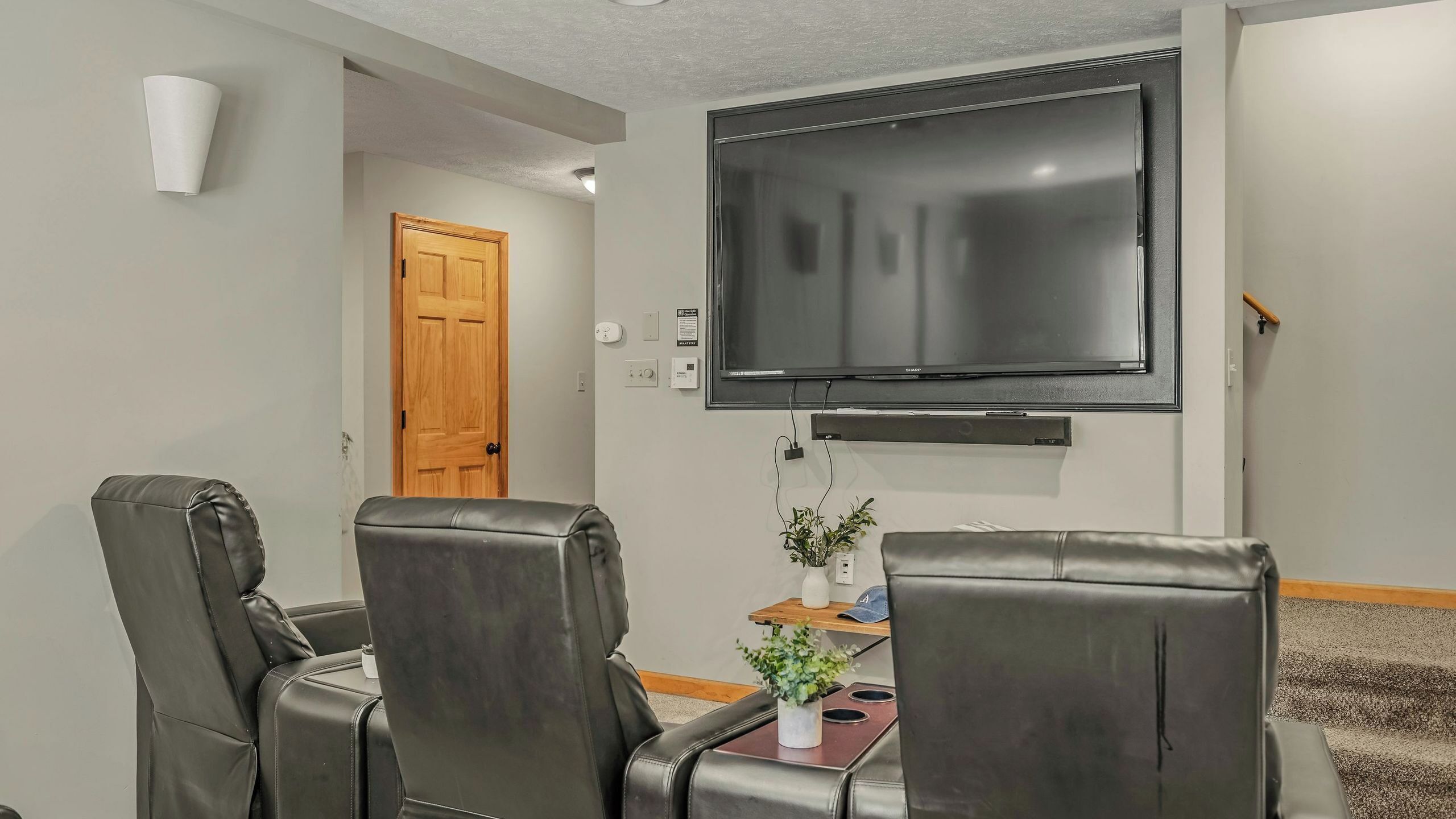 Un salon avec un téléviseur à écran plat fixé au mur, des fauteuils en cuir, une petite table avec une plante et une porte en bois.