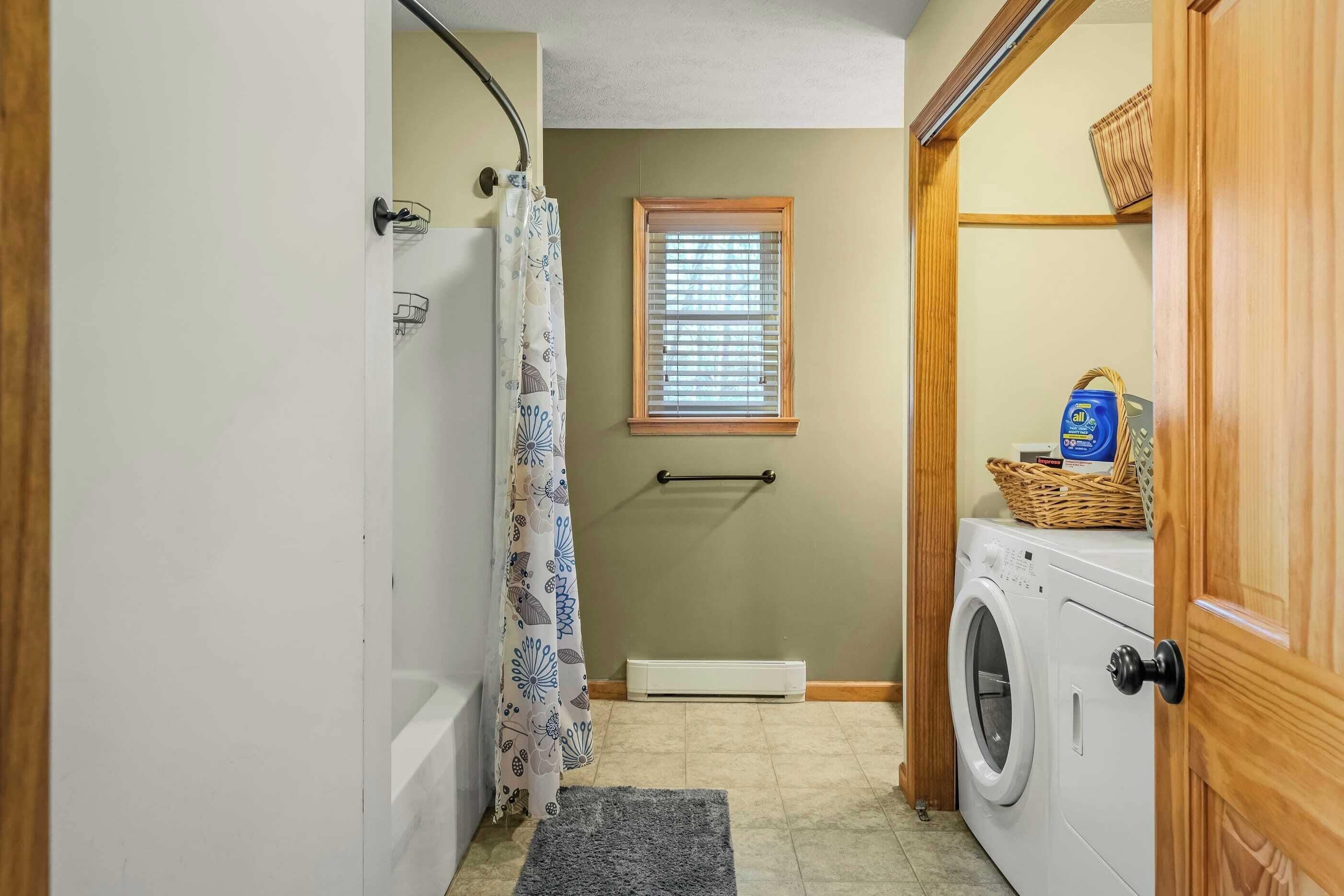 A laundry room with a washing machine, a drying rack, a window with blinds, and a shower curtain.
