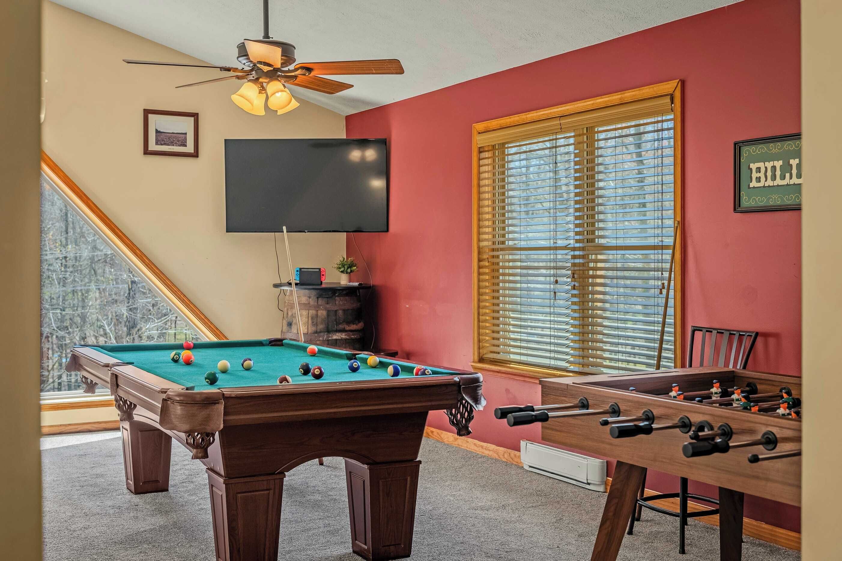 A pool table, foosball table, and a television in a room with a red wall and a window with blinds.