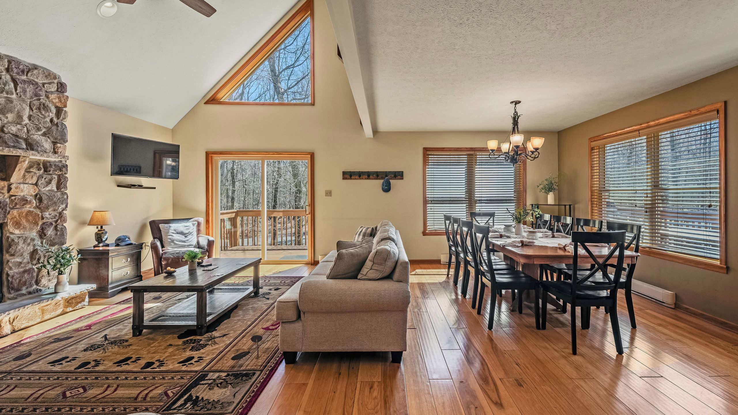 A spacious living room with a stone fireplace, a large sofa, a dining area with a wooden table and chairs, and large windows with blinds.