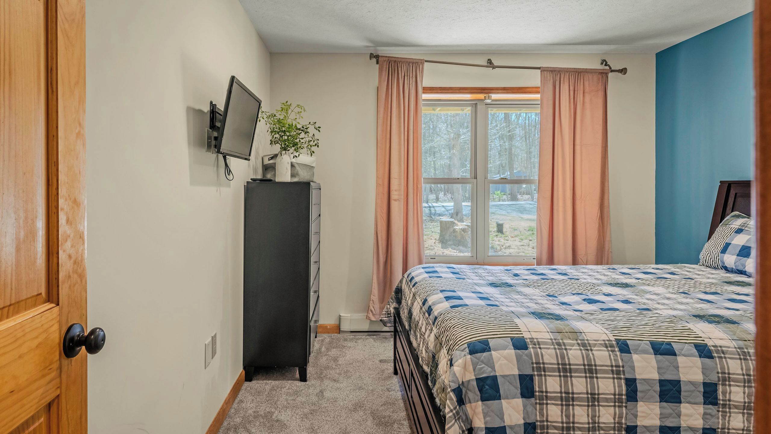 Une chambre à coucher avec un lit, une commode, un téléviseur fixé au mur et une fenêtre avec des rideaux.
