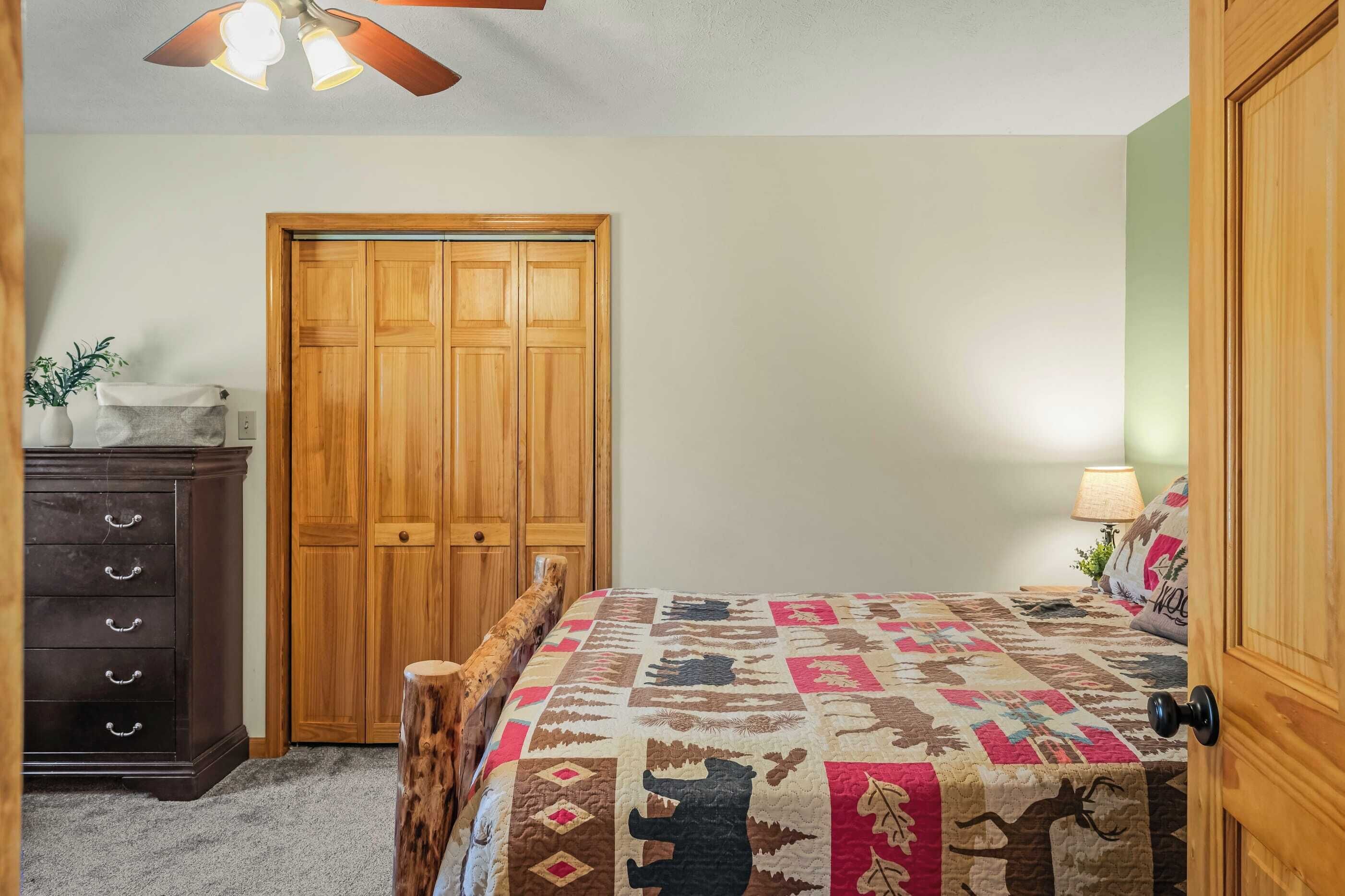 A bedroom with a wooden wardrobe, a bed with a patterned quilt, a nightstand with a lamp, and a ceiling fan.