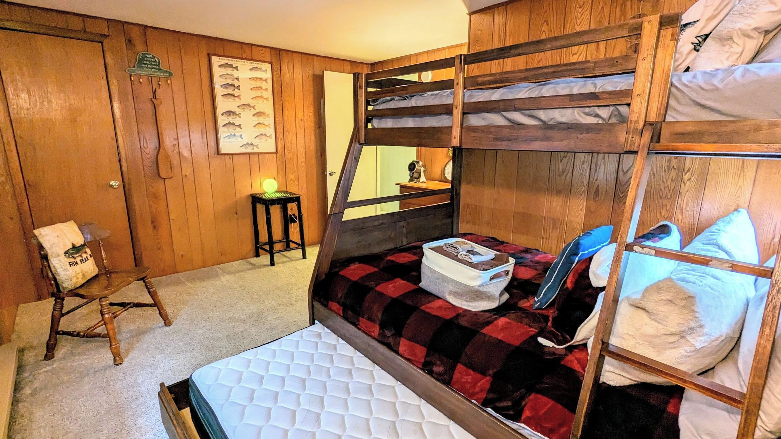 A bunk bed room with a wooden wall, a small stool, a framed fish chart, and a mirror.