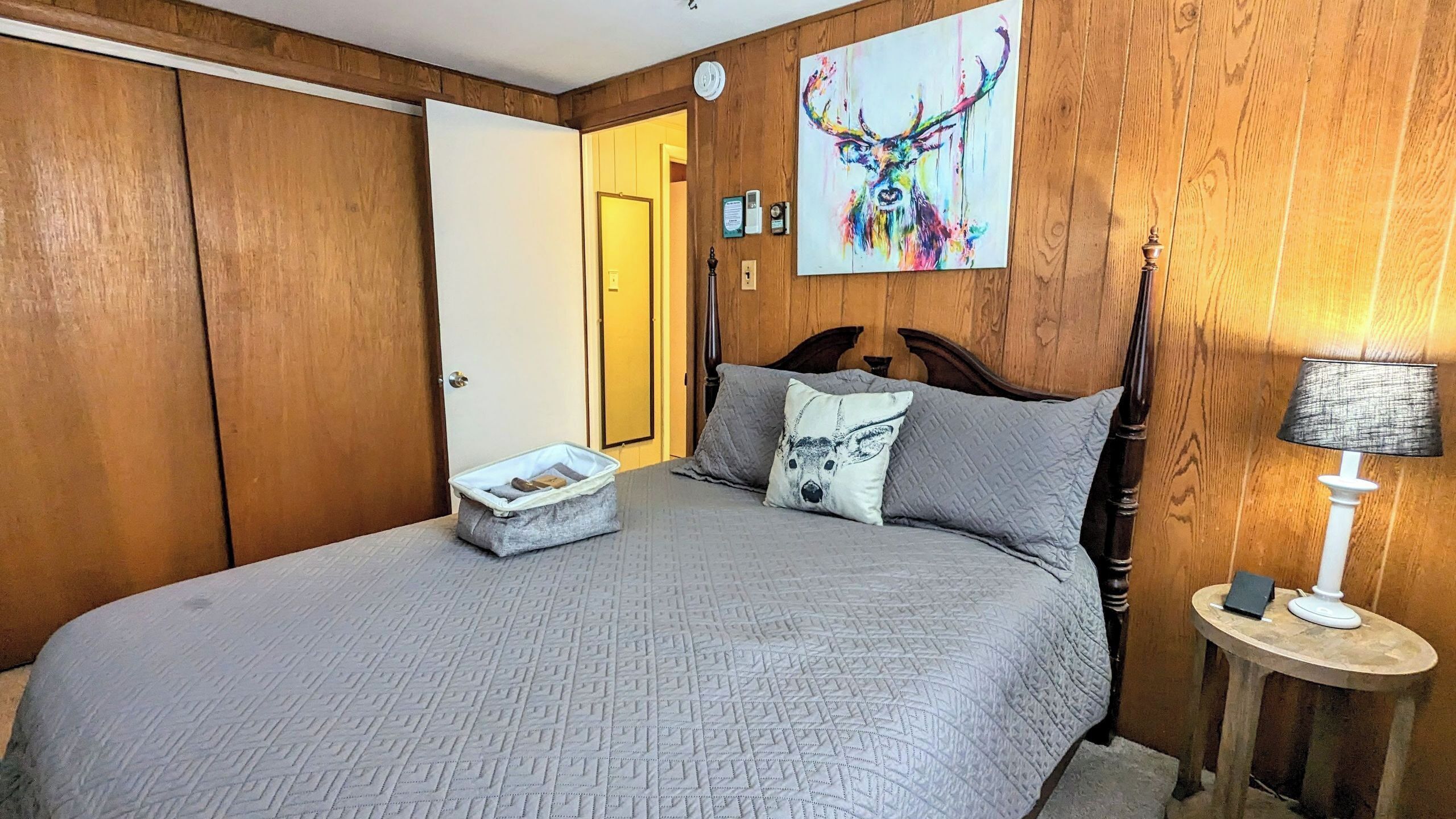 Un dormitorio con cama, mesita de noche, lámpara, puerta y un cuadro en la pared.