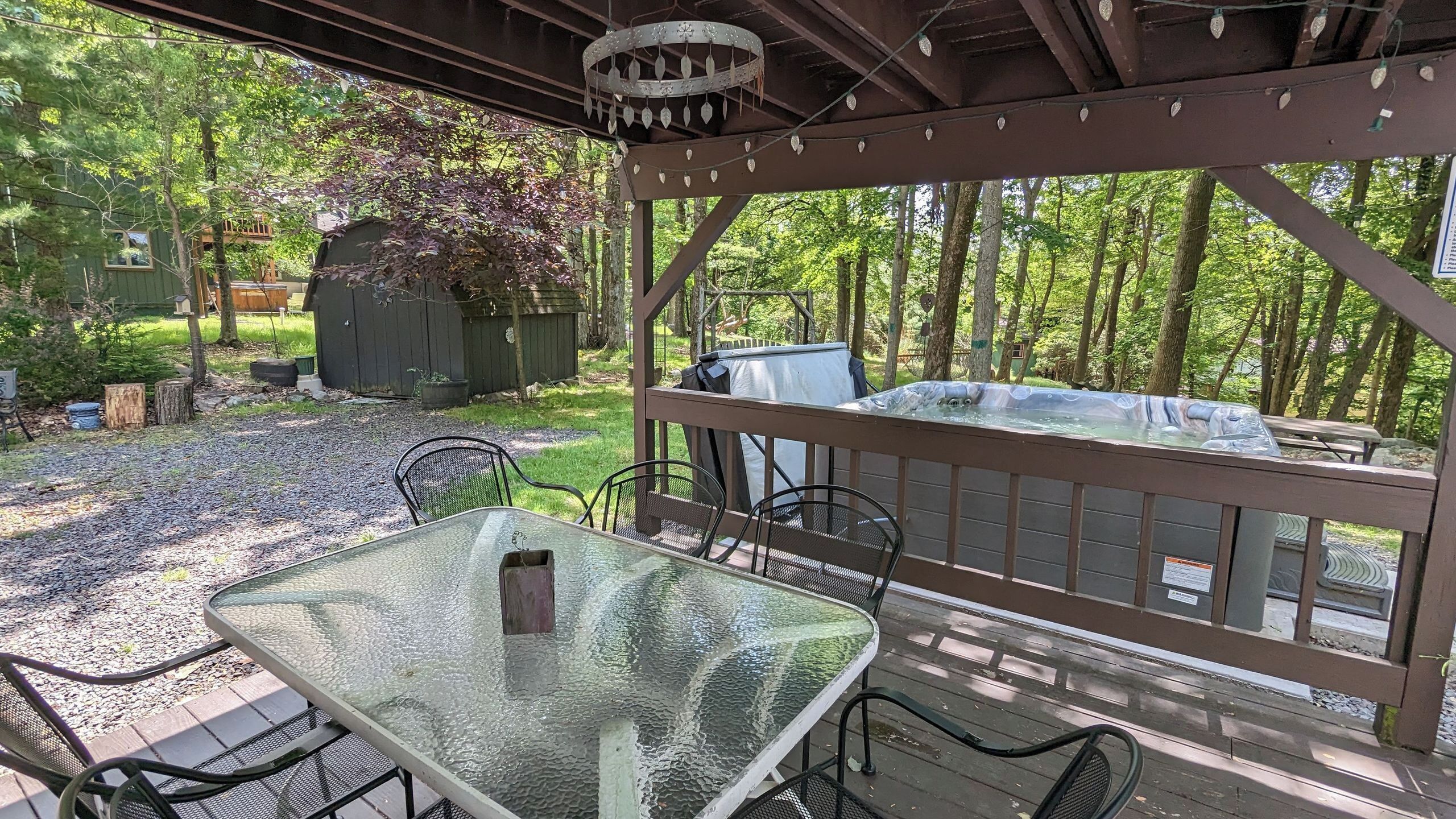 A covered outdoor seating area with a glass table and chairs, a hot tub, and a wooden deck.