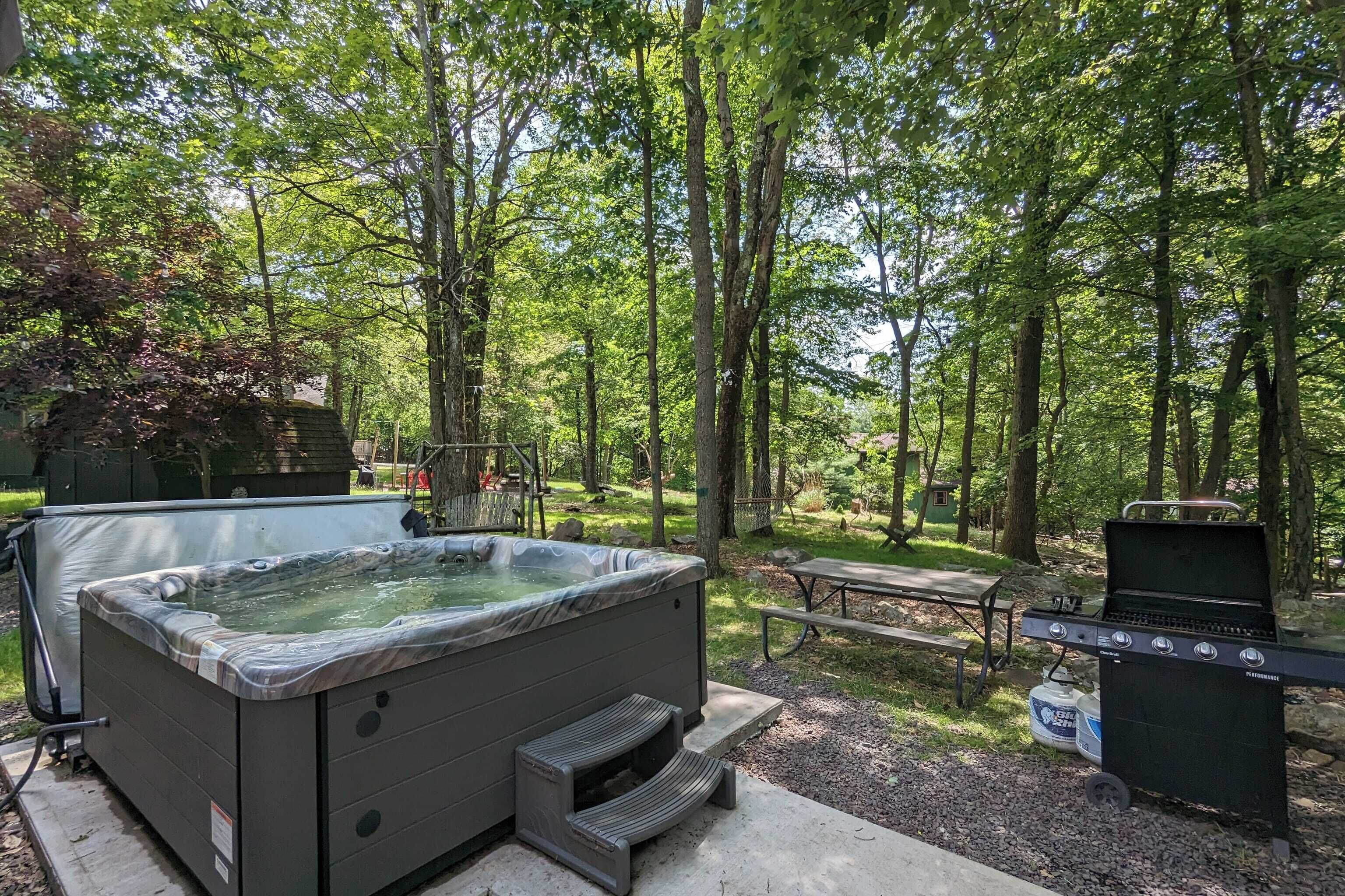 A hot tub, grill, picnic table, and a covered area in a wooded setting.
