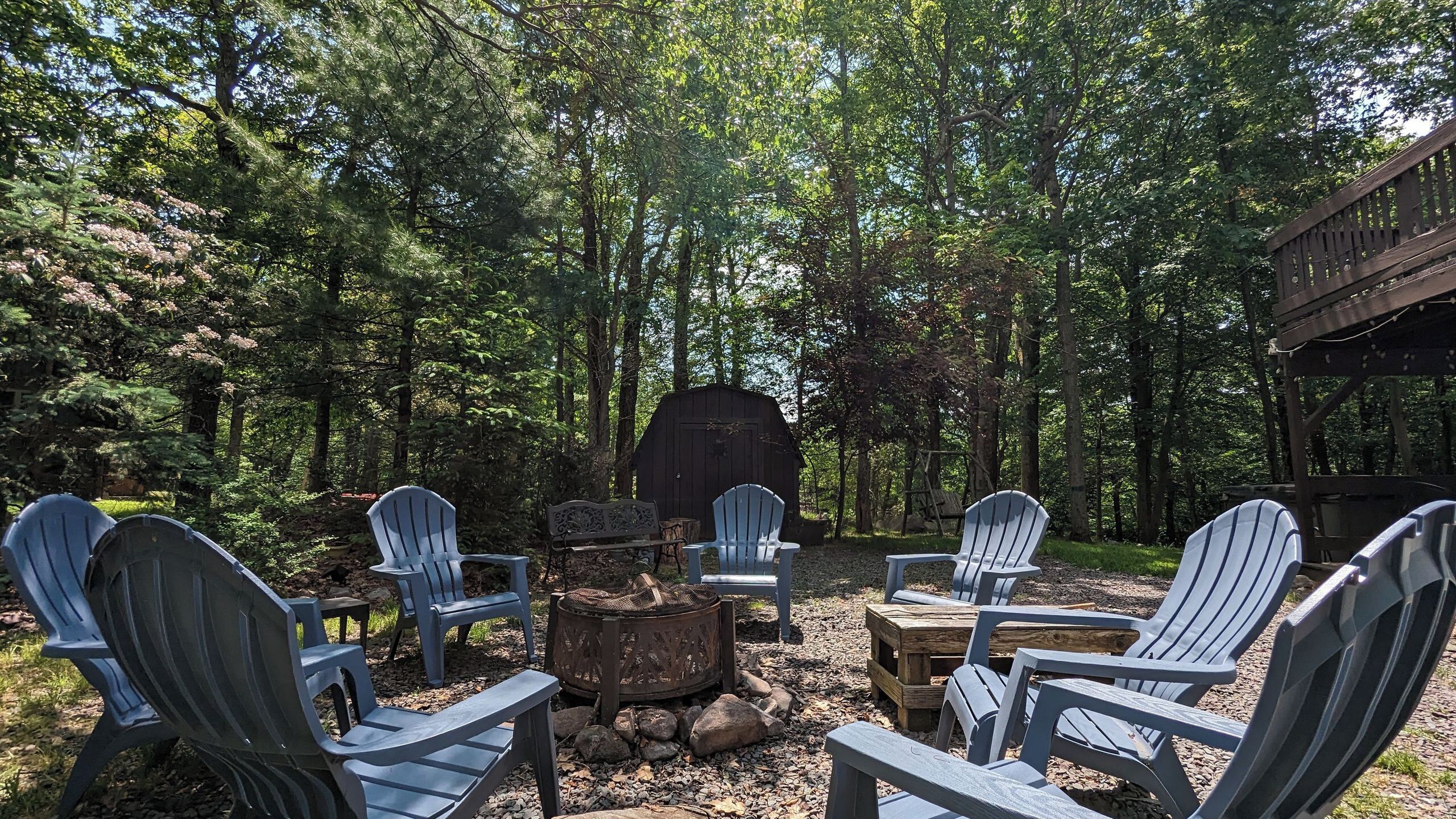 Een terras omgeven door bossen, met Adirondack-stoelen, een vuurkorf en een houten constructie.