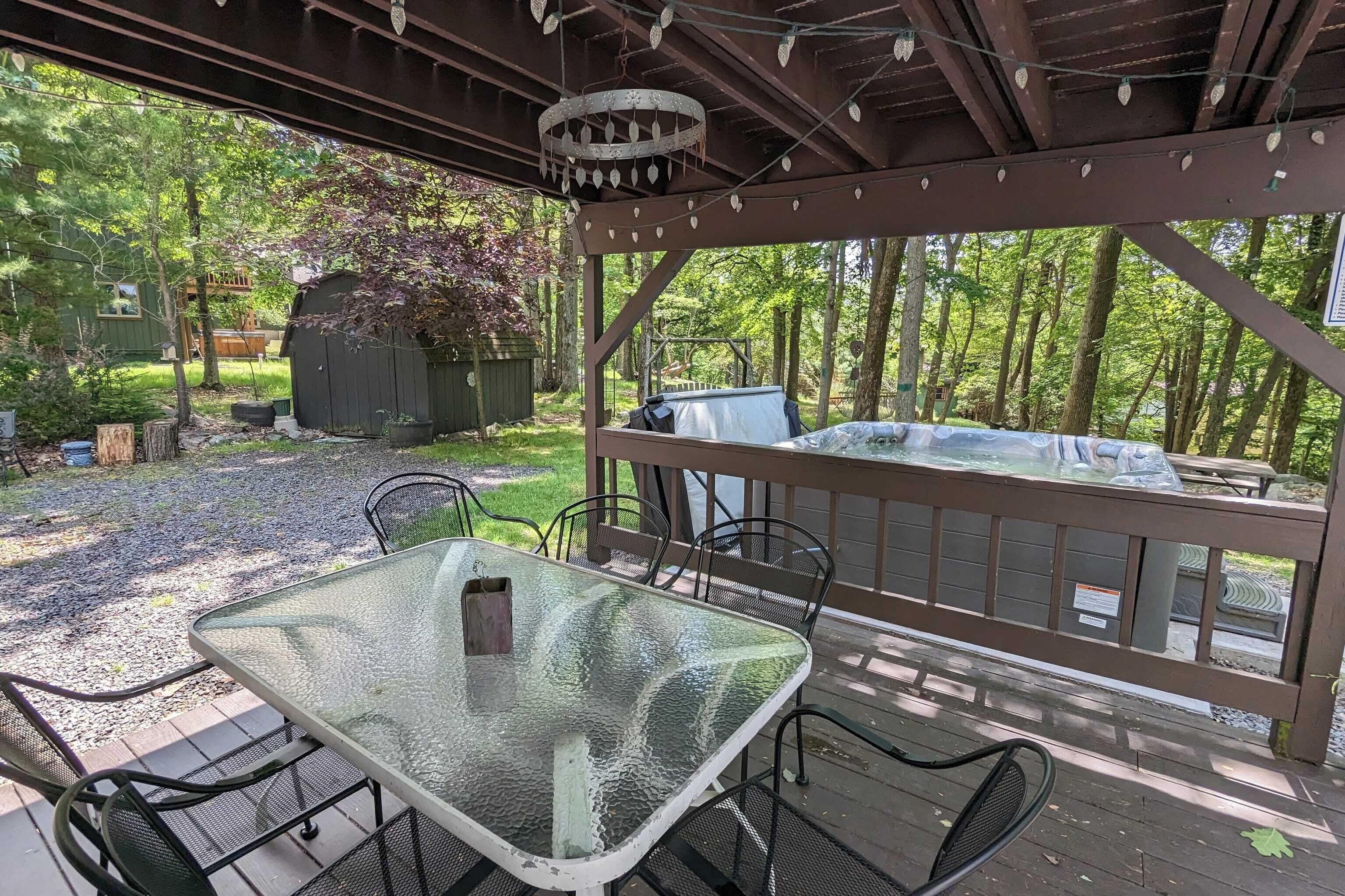 En overdækket terrasse med et glasbord og stole, omgivet af et træterrasse og udsigt til et skovområde.