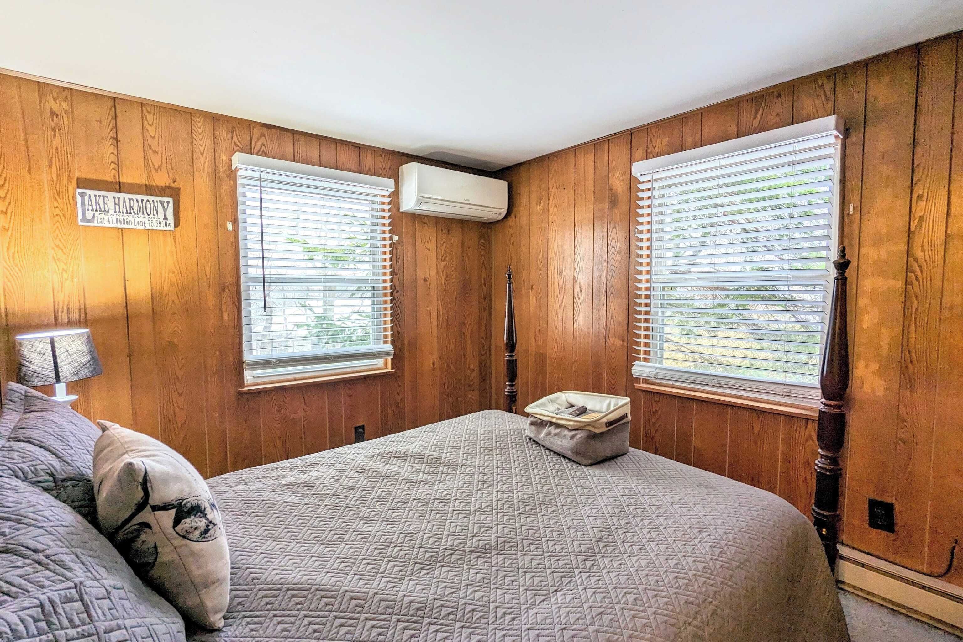 A bedroom with wooden walls, a bed with a quilted bedspread, two windows with blinds, an air conditioning unit, and a small bedside table with a lamp.