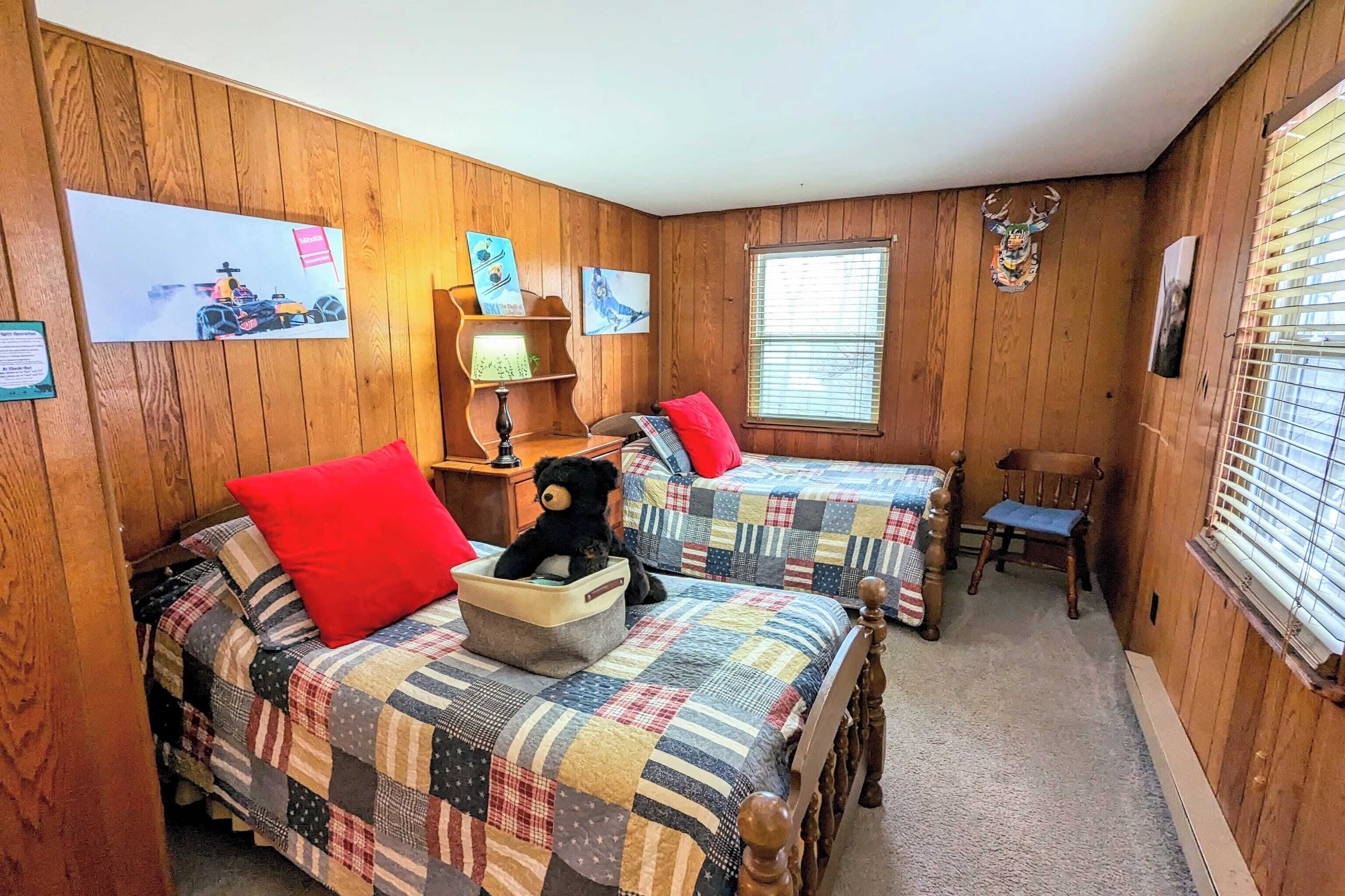 A cozy room with wooden walls, two beds with checkered quilts, a red pillow, a teddy bear, a small table with a lamp, and a window with blinds.