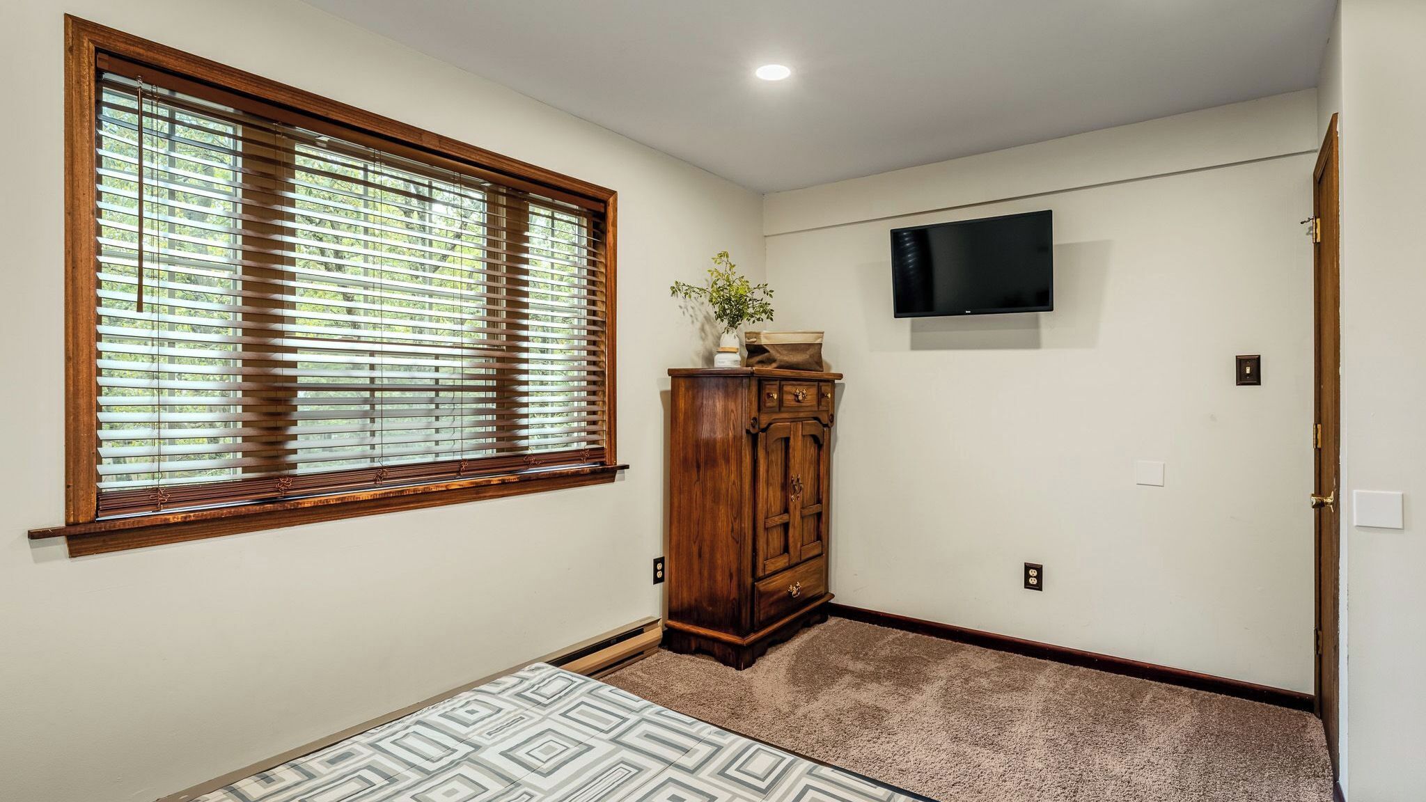 A room with a large window, a wooden cabinet, a television on the wall, and a patterned rug.