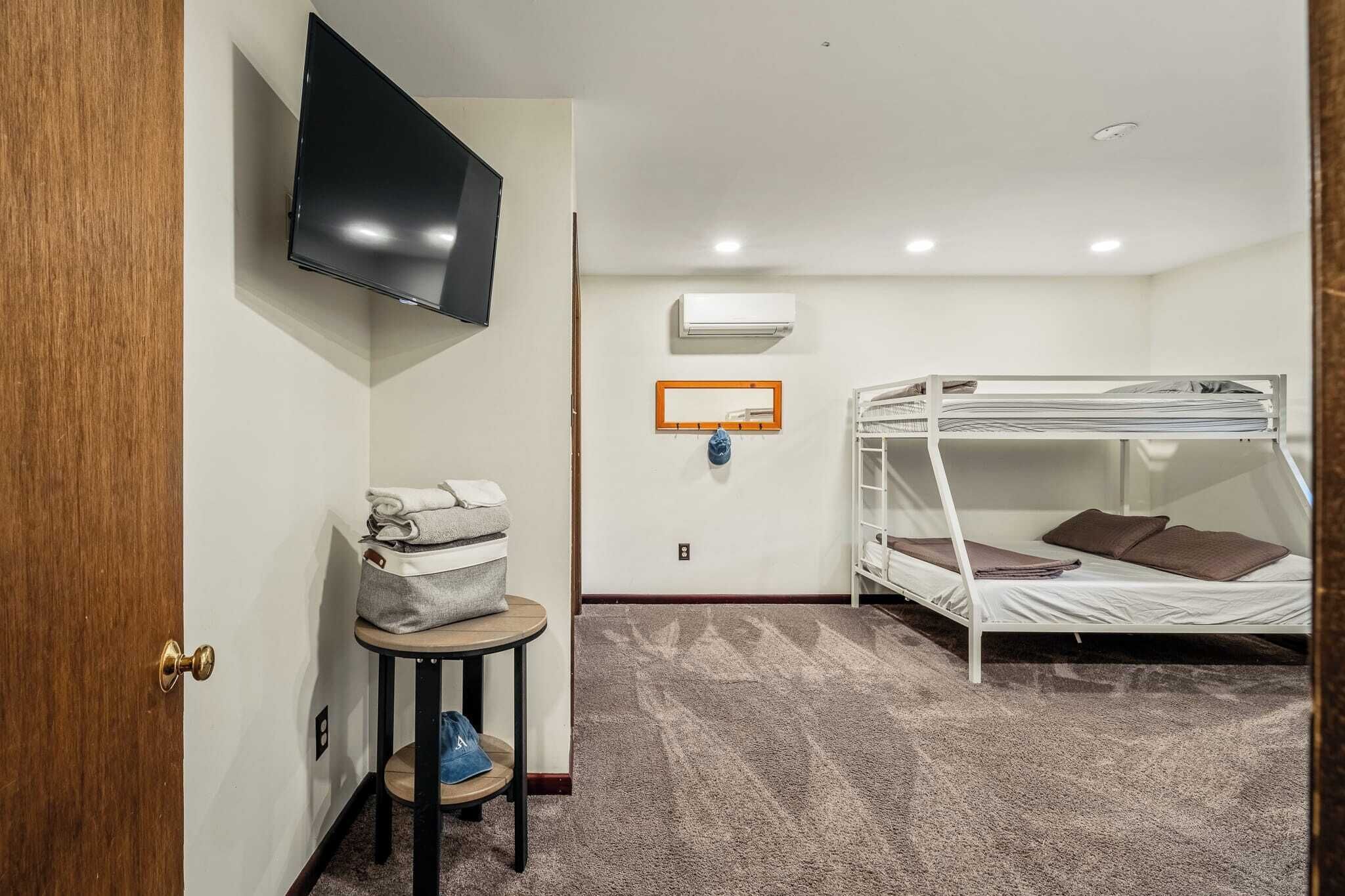 A room with a bunk bed, a television mounted on the wall, a small table with a bag and a blue item, and an air conditioning unit.