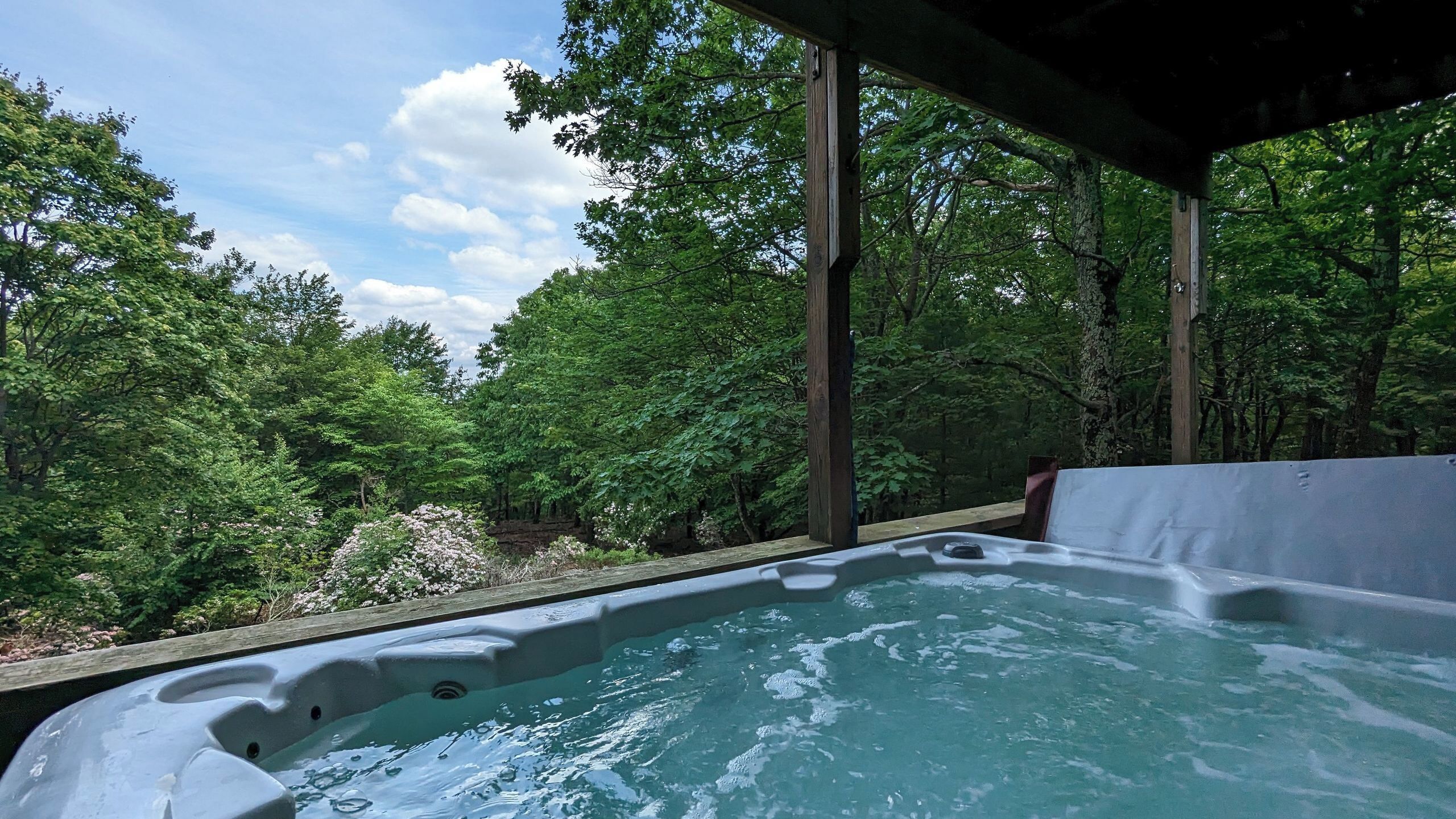 Een hottub in een bebost gebied, met een houten terras en een overdekt gedeelte.