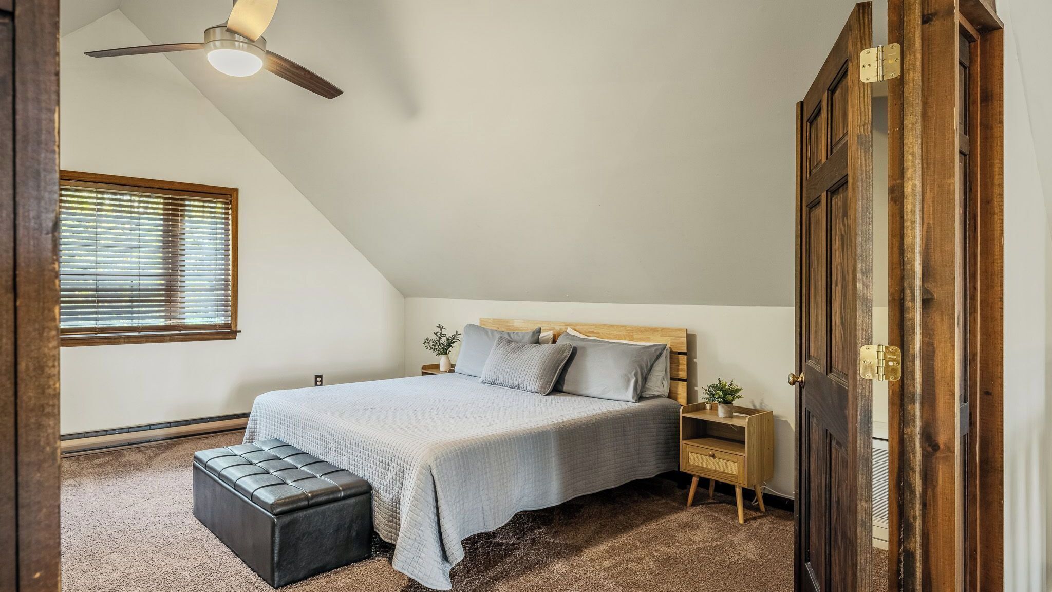 Une chambre avec un plafond en pente, un lit doté d’une tête de lit en bois, un banc, une table de chevet, une plante en pot et une porte avec un cadre en bois.