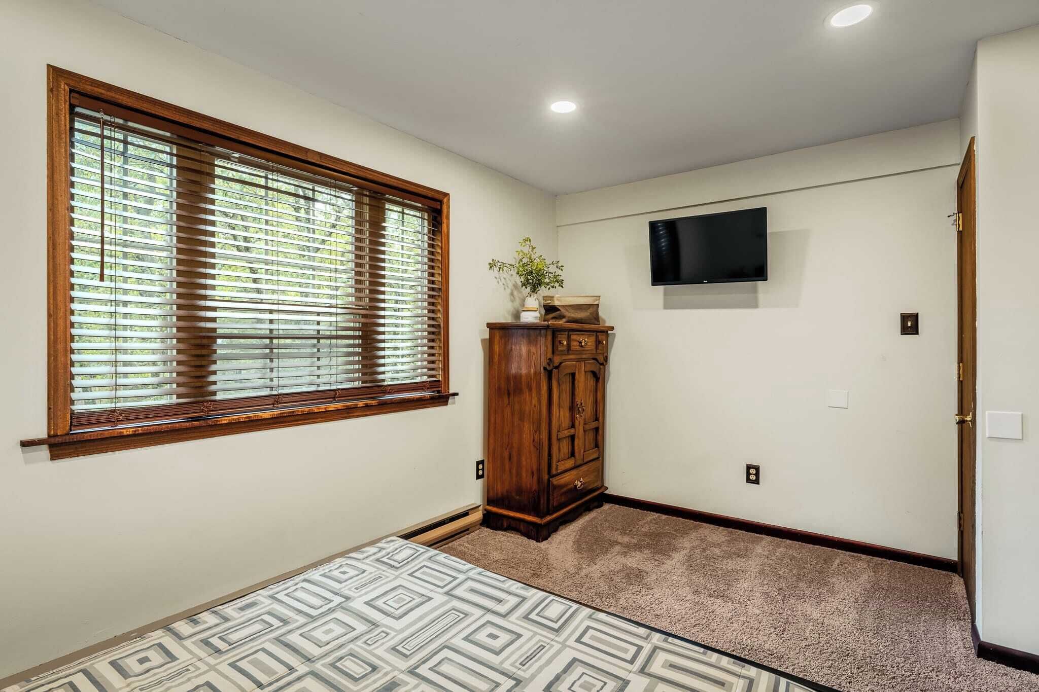 A room with a large window with blinds, a wooden cabinet, a flat-screen TV mounted on the wall, and a patterned carpet.