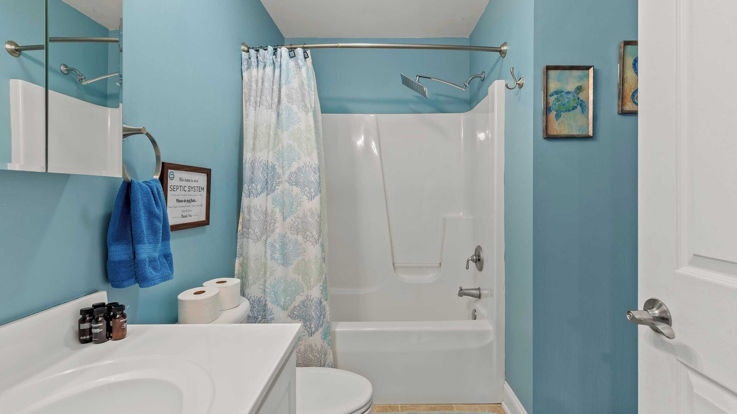 Un baño con bañera, ducha e inodoro. Hay un perchero con una toalla azul, un cartel enmarcado y algunas botellas sobre el lavabo.