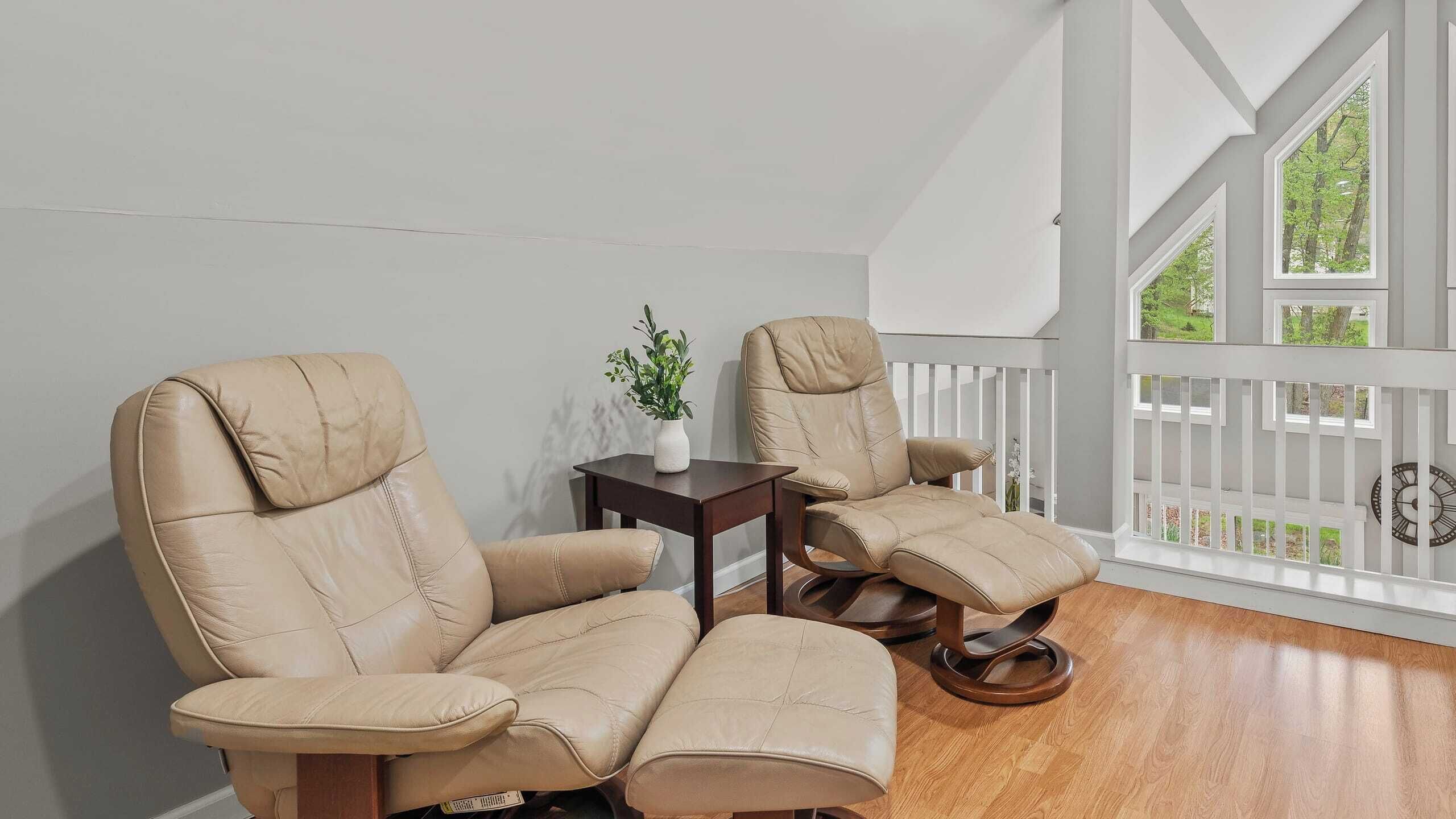 Una habitación con dos sillones de cuero color beige, una mesita de madera con un florero de plantas verdes, y vistas a una cerca blanca y follaje exterior.