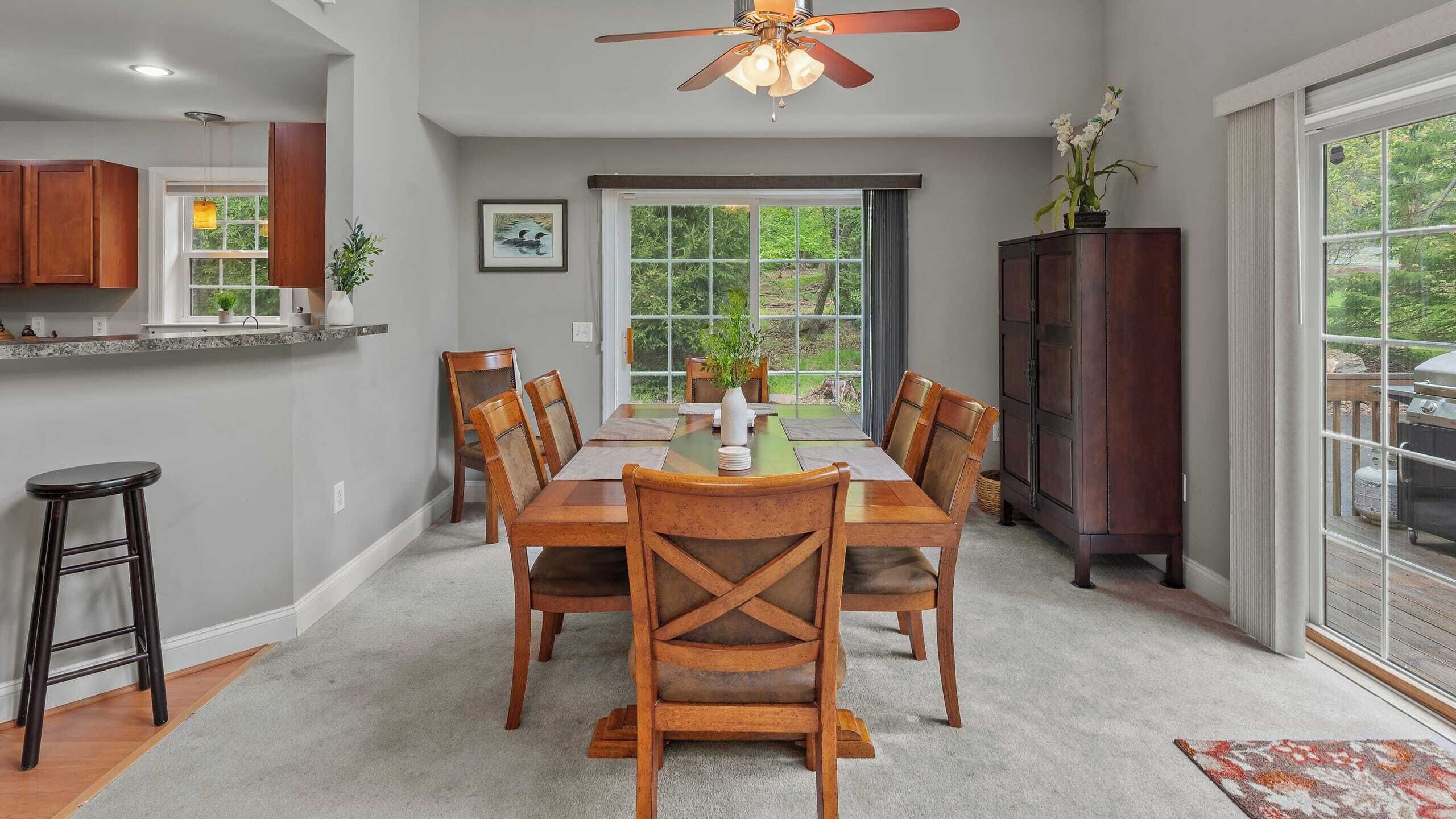 Un espace repas avec une table et des chaises en bois, un ventilateur de plafond et une vue sur l’extérieur grâce à de grandes fenêtres.