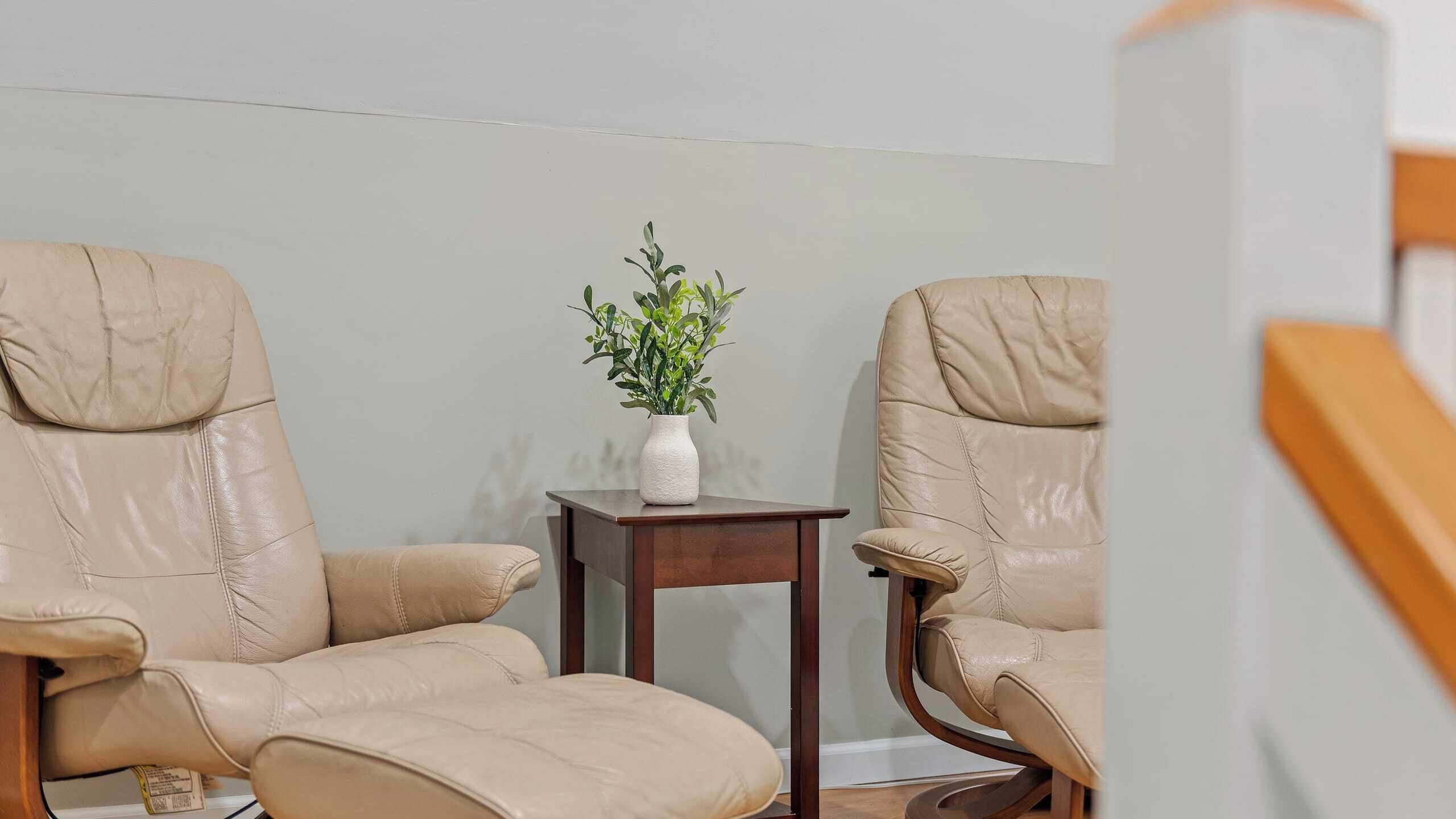 Une pièce d’angle avec deux fauteuils en cuir, une petite table d’appoint en bois sur laquelle se trouve un vase blanc contenant une plante, et un mur uni de couleur claire.