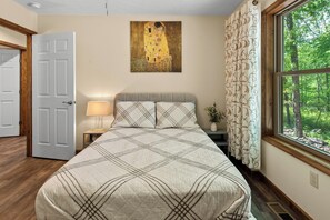 A bedroom with a bed, a nightstand, a lamp, a painting, a window with a forest view, and a door.