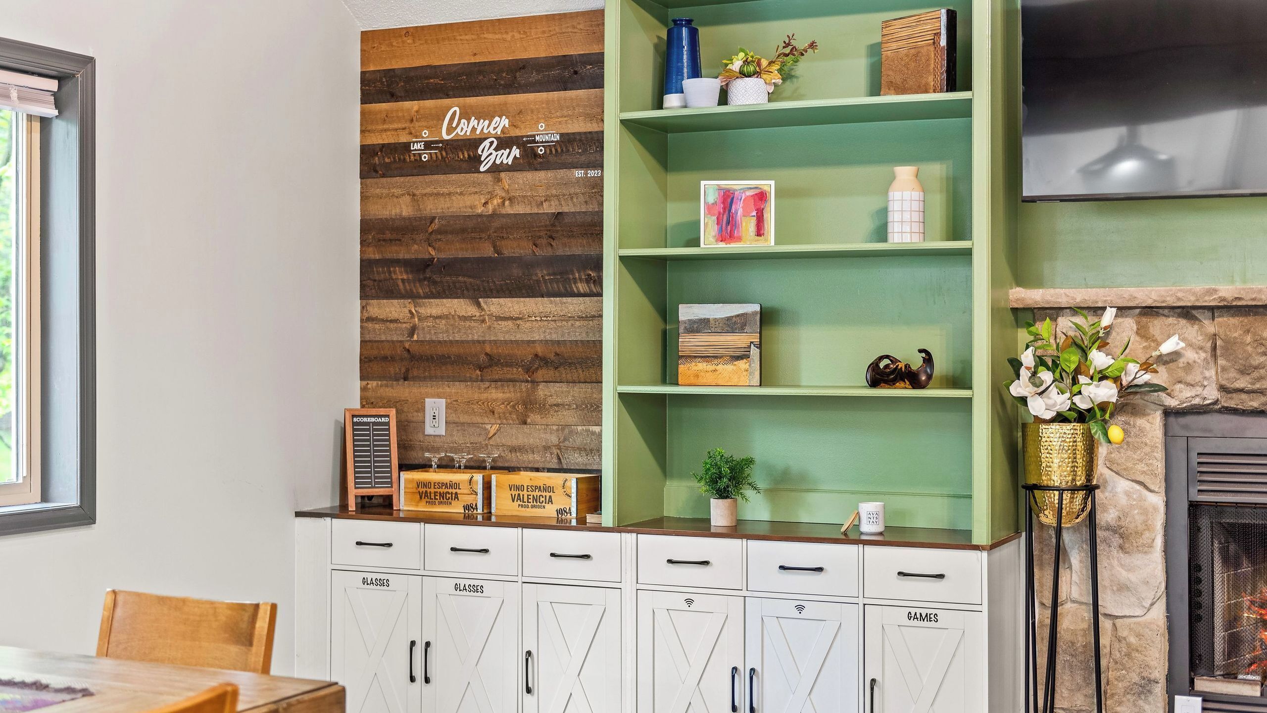 Ein Zimmer mit einer Holzwand, einer grünen Regaleinheit mit Dekoration, einem Kamin und weißen Schränken, auf denen 'GLÄSER' und 'SPIELE' steht.