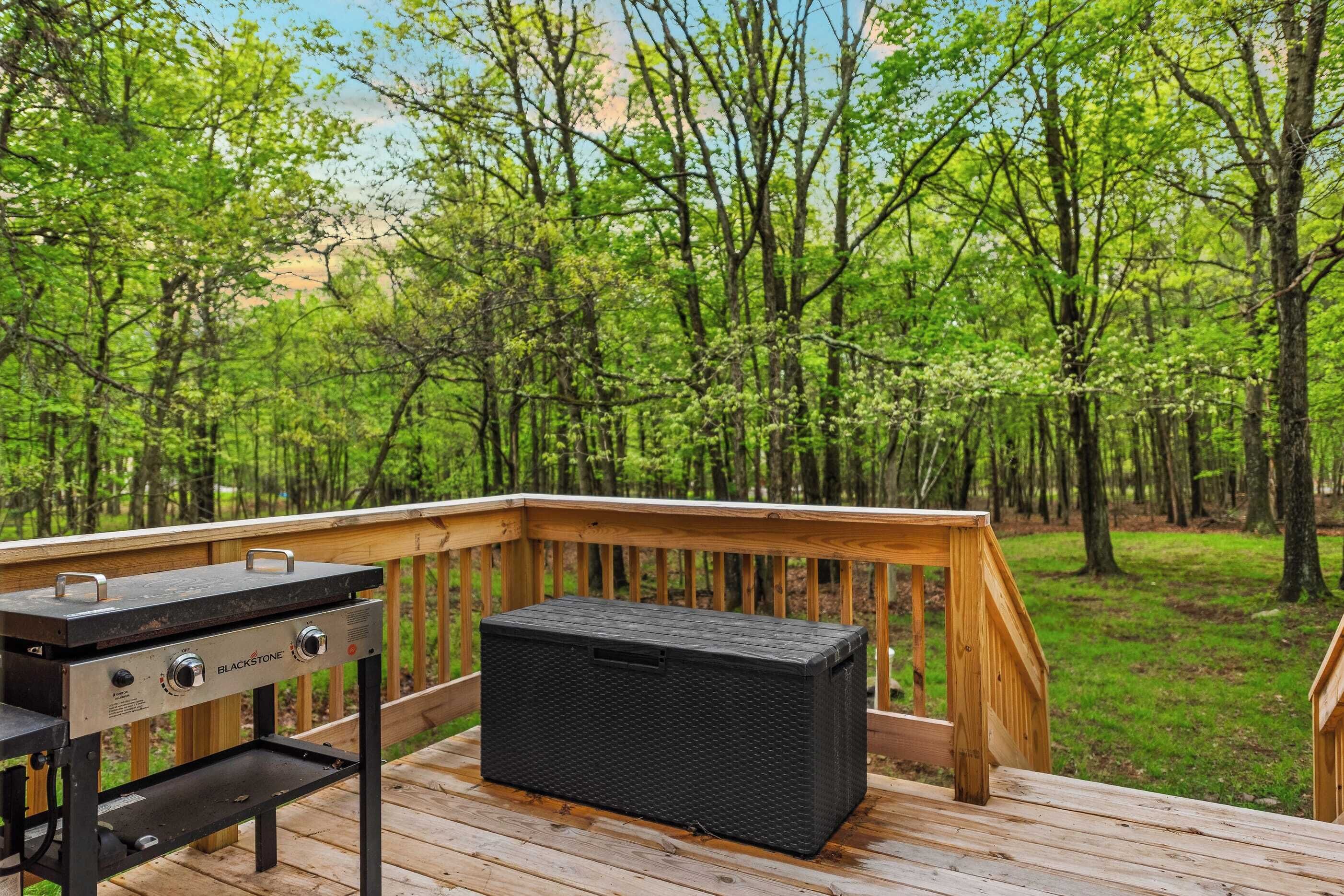 A deck with a Blackstone grill and a storage box, surrounded by a forested area.