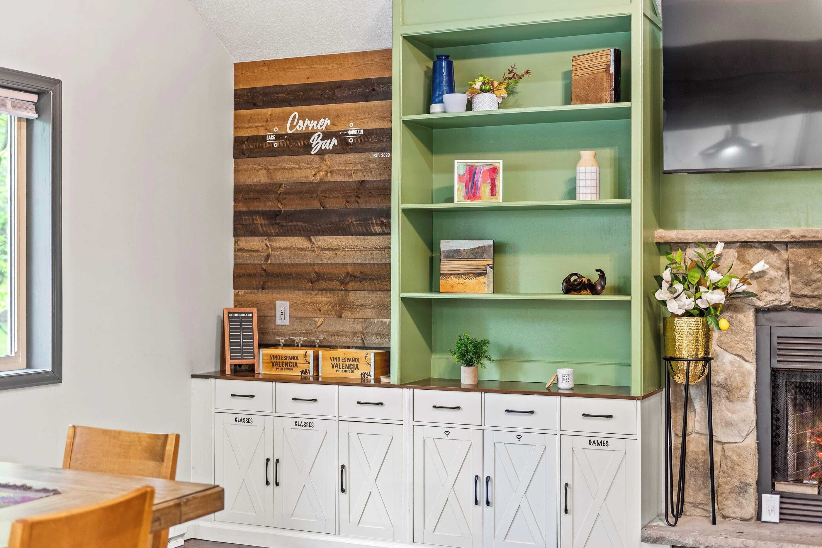 Un rincón de bar acogedor con pared de madera, estanterías verdes y armarios blancos con la etiqueta 'GLASSES' y 'GAMES'.