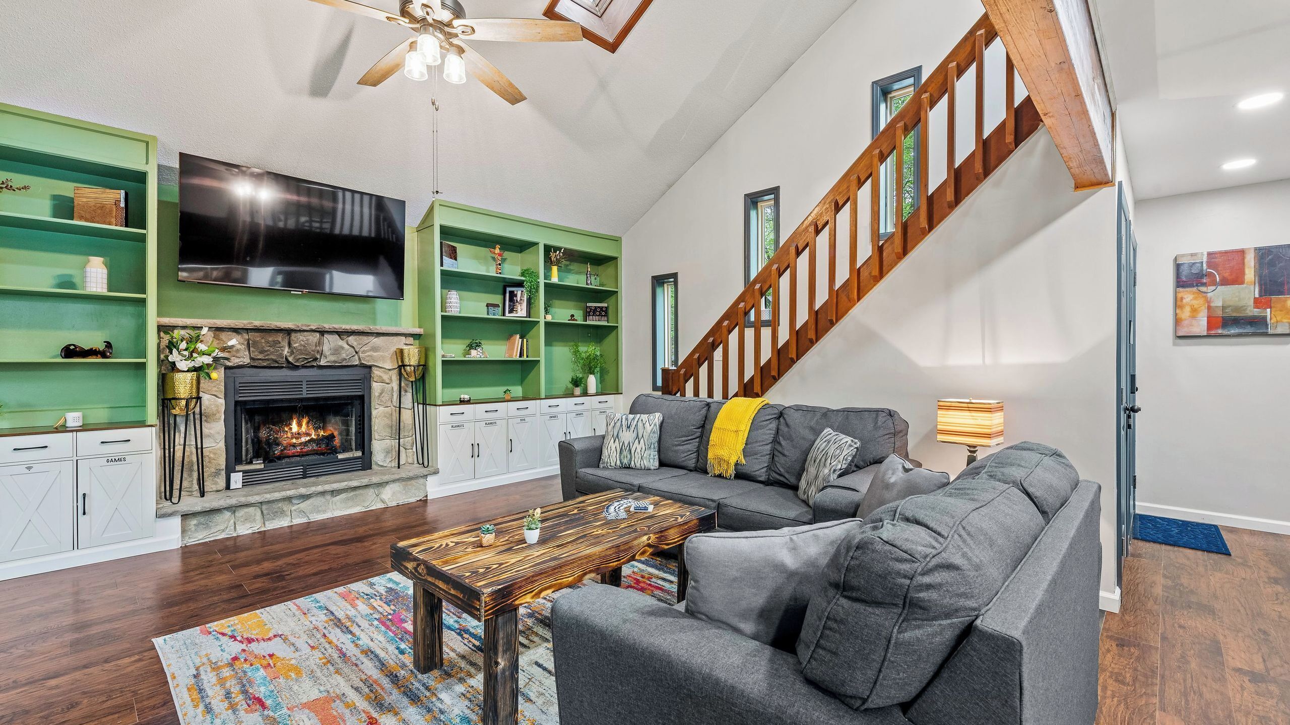 A living room with a fireplace, green shelves, a gray sofa, and a wooden staircase.