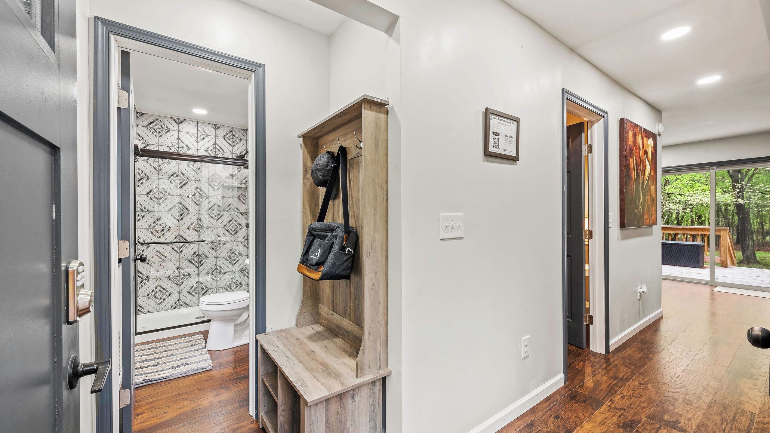 A hallway with a wooden bench, a door leading to a bathroom with patterned tiles, and a view of a deck with trees.