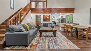 A spacious living area with a staircase leading to an upper level, a dining area with wooden furniture, and a kitchen in the background. - The Tranquil Trails by Avantstay Lake Access, Deck, Fireplace (Albrightsville)