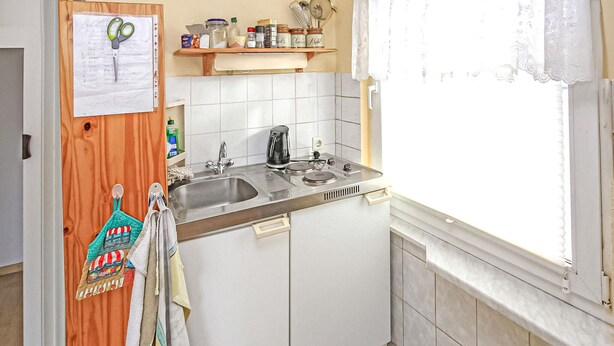 Pieni keittiö, jossa on pesuallas, liesi ja ikkuna. Puuovessa roikkuu paperi ja parit sakset. Näkyvissä on myös pyyheteline, jossa on värikäs pyyhe.