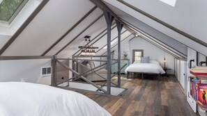 Une chambre à coucher aménagée dans un espace mansardé, avec un plafond en pente, des poutres apparentes, un lit, un bureau et une bibliothèque.