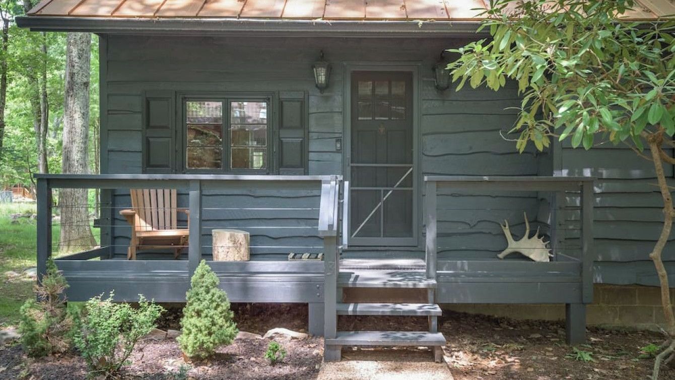 Een houten hutje met een veranda, een stoel en een kleine boom in de voortuin.